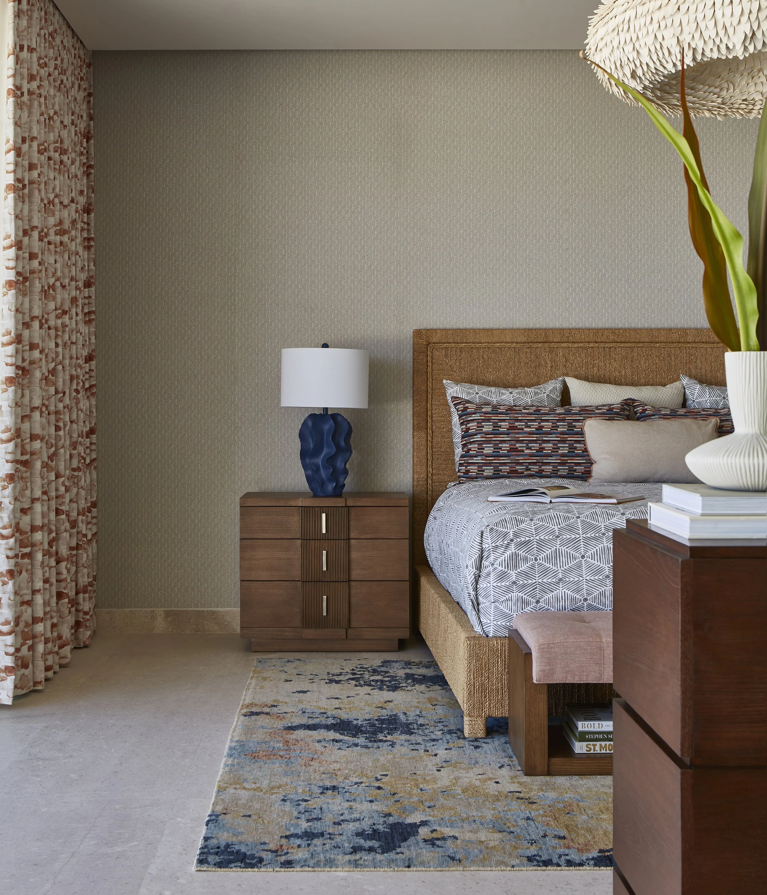 A bedroom with a wooden bed, patterned bedding, a bedside table with a blue lamp, colorful curtains, a rug, and a large white vase with a green plant.