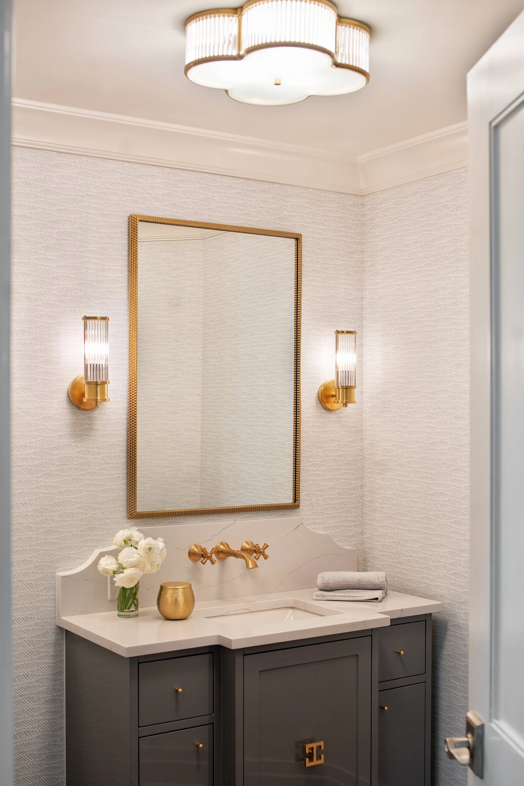 Bathroom with a gold-framed mirror, gold wall sconces, a gray vanity, white flowers in a vase, a gold container, a gray towel, and a white marble countertop.