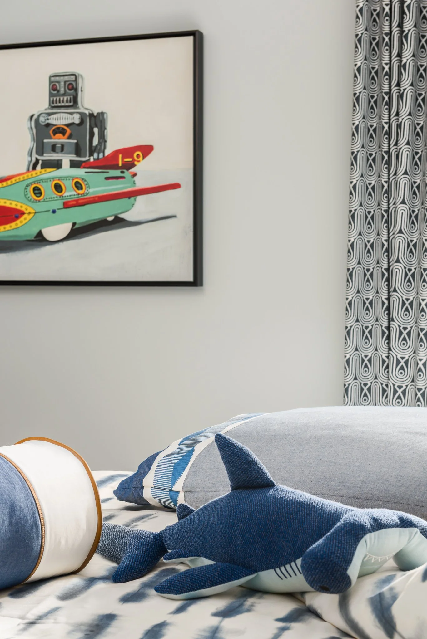 A bedroom with a bed that has a shark-shaped plush toy and a white cylindrical pillow with a brown trim. There is a framed picture of a toy robot and a colorful toy submarine on the wall, and blue-coordinated bedding and curtains with a geometric pat