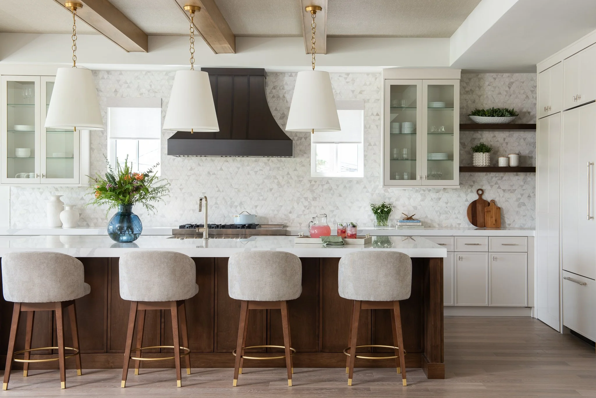 Modern kitchen with a white island, beige chairs, wooden ceiling beams, open shelving with white dishes, and a mosaic backsplash.