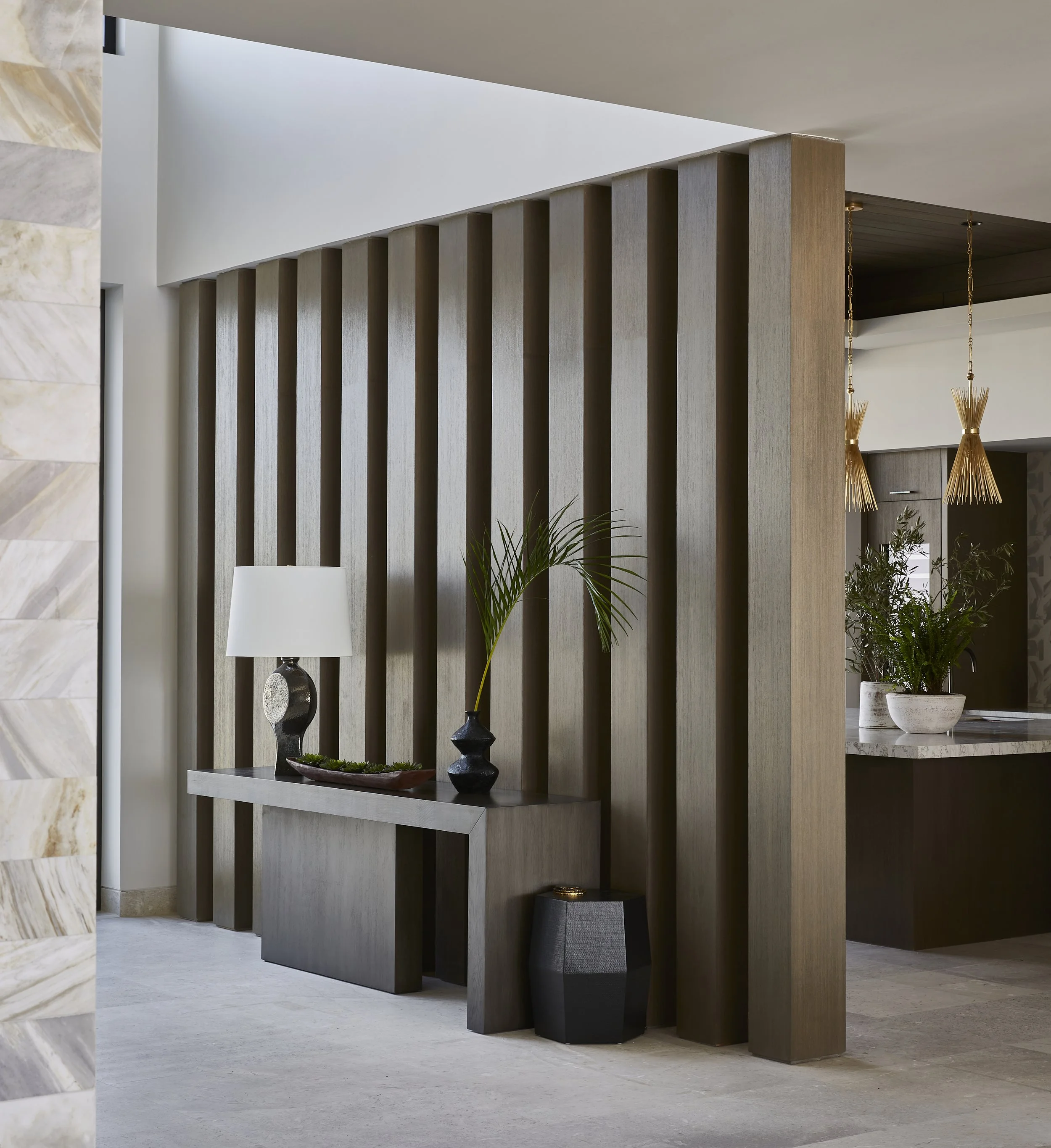 Modern interior with a wooden slat partition, a console table with a table lamp, vase, and decorative bowl, and potted plants in the background.