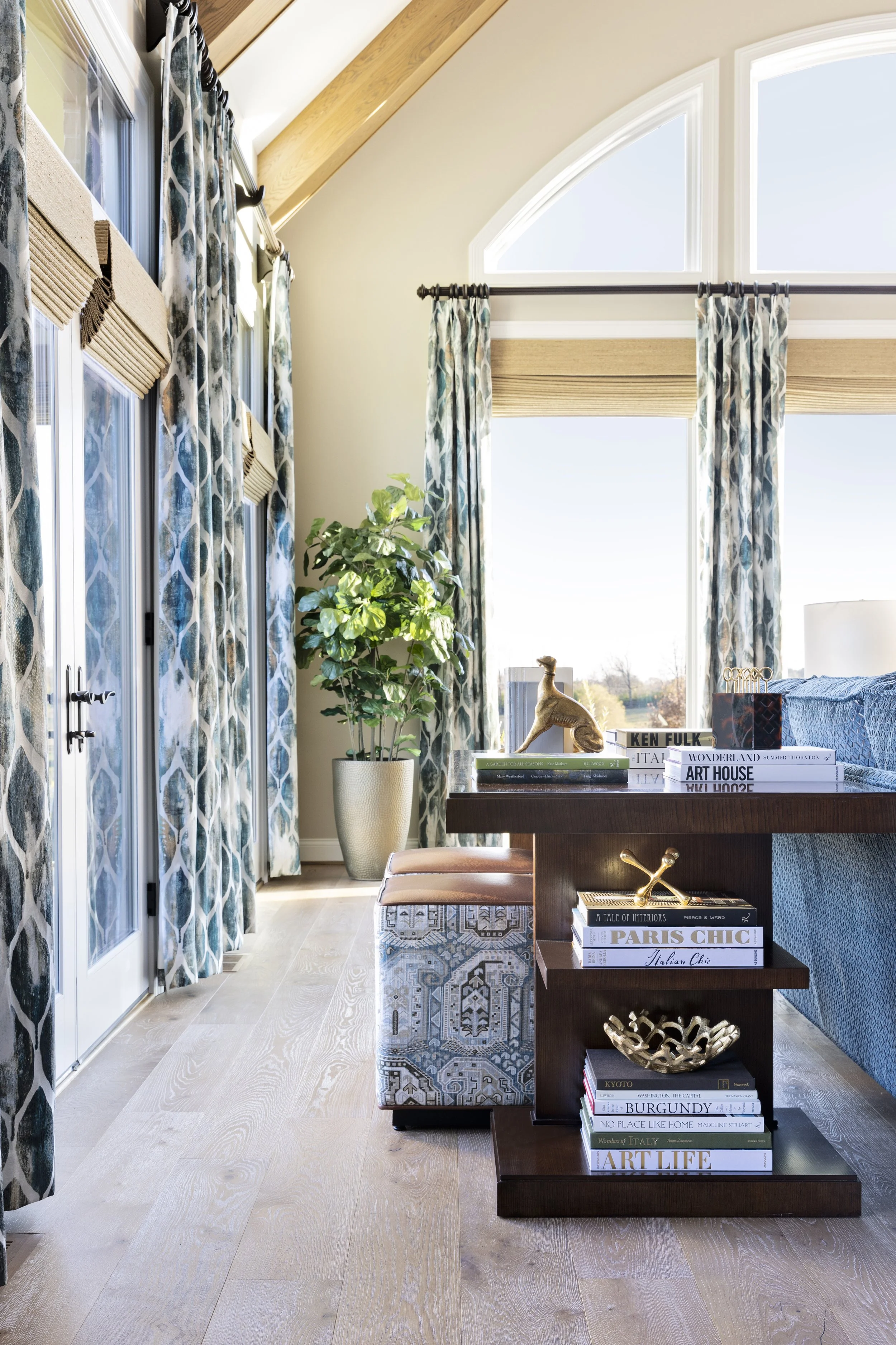 A bright living space with large windows and arched window above. Light-colored hardwood floors, patterned curtains, a large potted plant, and a wooden table with books, decorative objects, and a patterned ottoman.