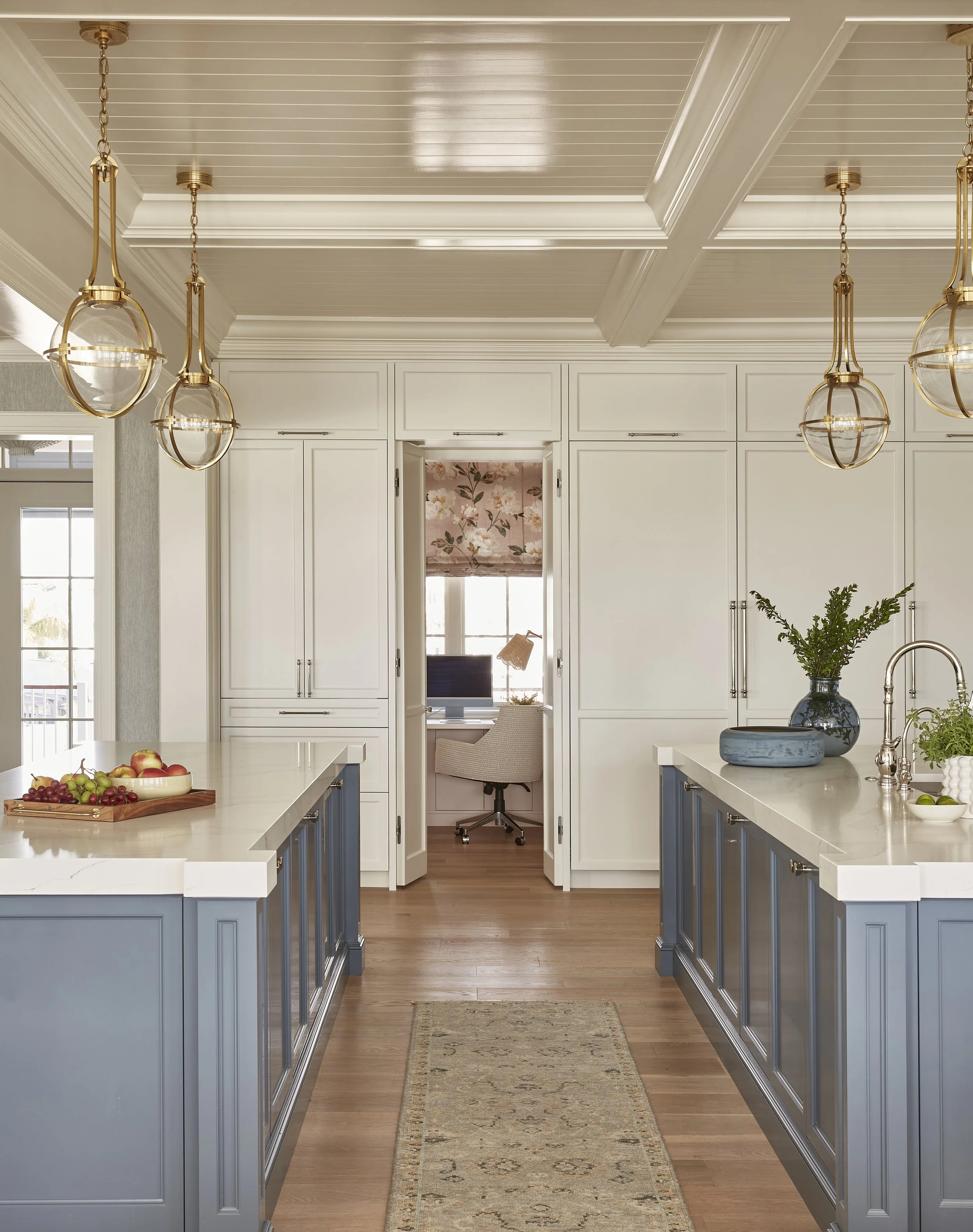 Modern kitchen with white cabinets, blue island cabinets, gold pendant lights, hardwood floors, and a view into a home office.