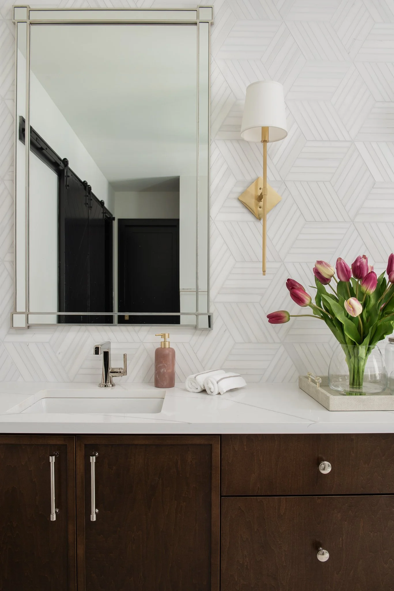 Modern bathroom vanity with dark wood cabinets, white countertop, a sink, a gold and white wall sconce, a large mirror, a pink soap dispenser, rolled white towels, and a glass vase with pink tulips.