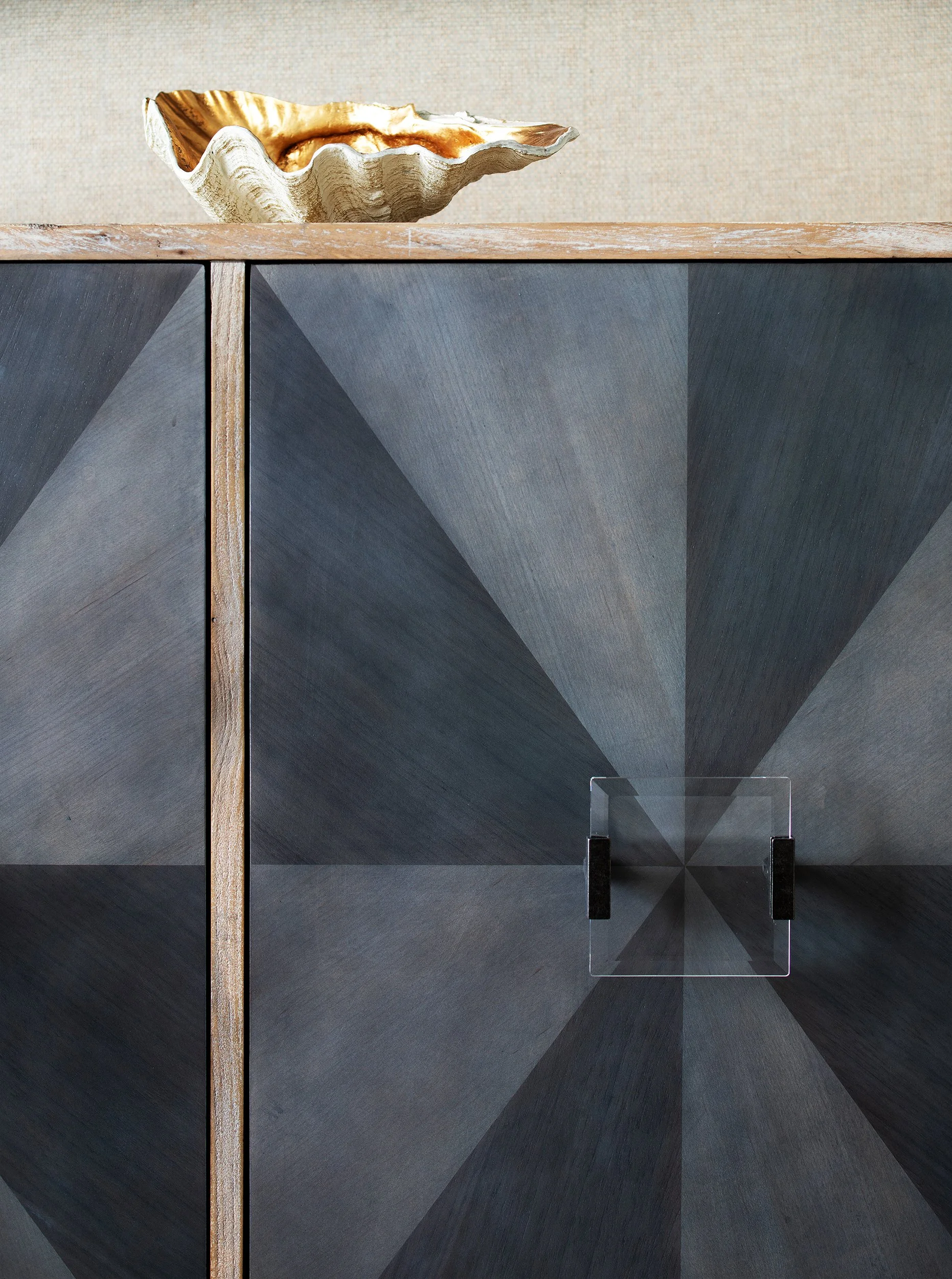 A decorative cabinet with a geometric dark gray pattern on its doors, featuring a black handle and a glass panel, topped with a large shell-shaped ceramic bowl with a gold interior on a wooden surface.