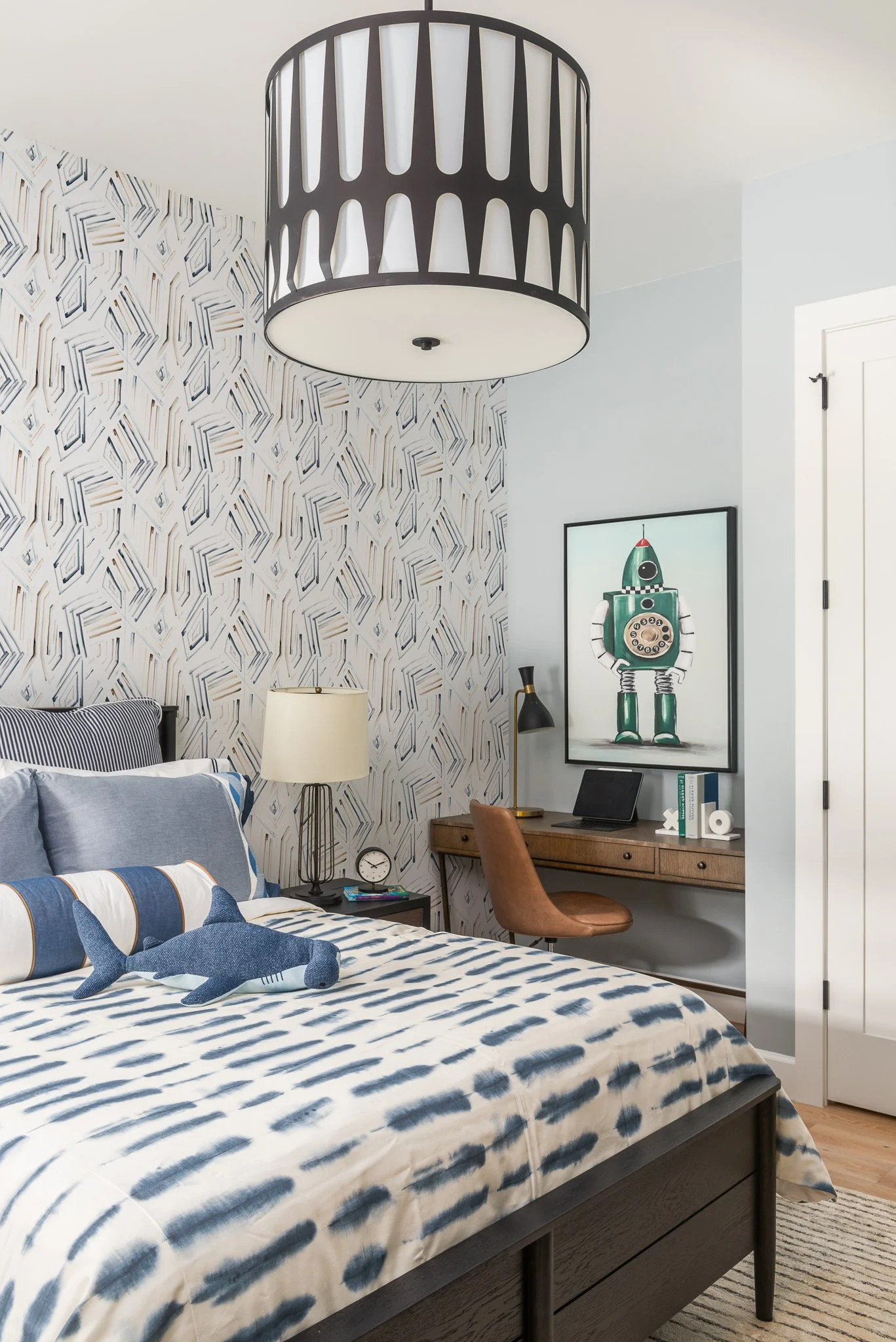 A bedroom with a bed featuring blue and white patterned bedding, a plush dolphin toy, a bedside lamp, a wall with an abstract geometric wallpaper, a small wooden desk with a leather chair, a framed robot poster, and a large drum shade ceiling light.