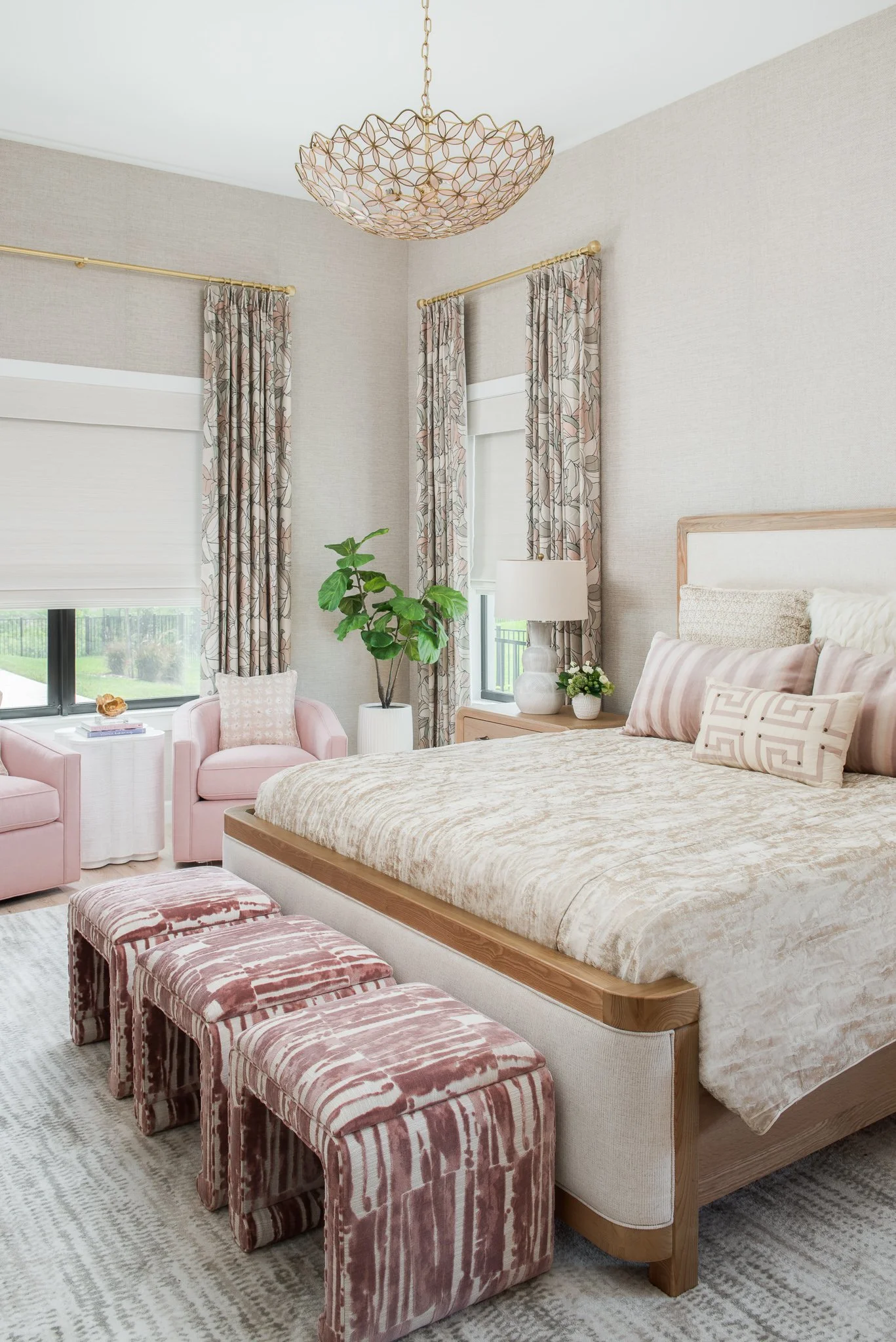 A bedroom with a large bed, pink patterned ottomans at the foot, a nightstand with a lamp, pink armchair, window with floral curtains, and a chandelier hanging from the ceiling.