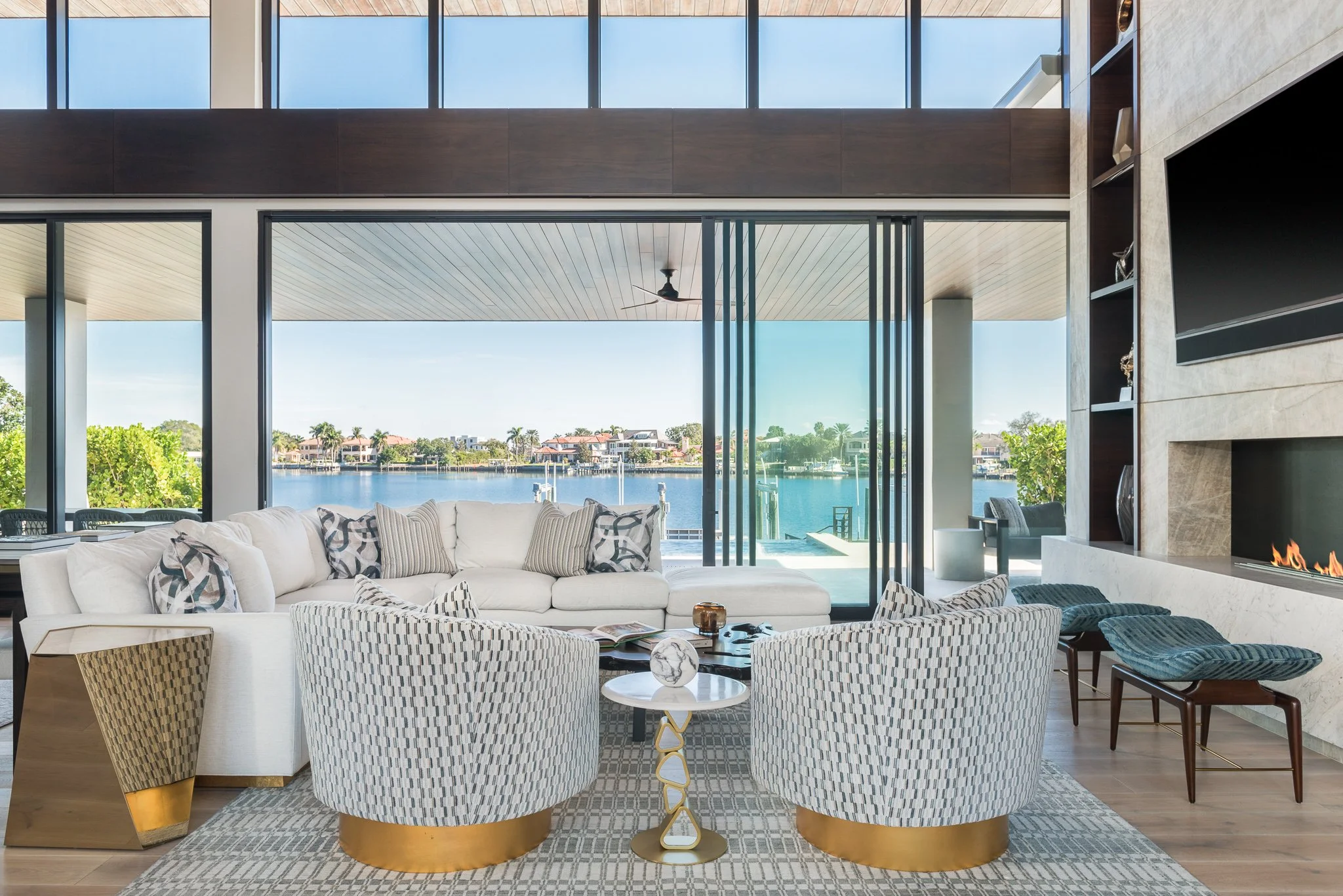 Modern living room overlooking a water view with large glass windows, a white sectional sofa, patterned armchairs, and a built-in fireplace.