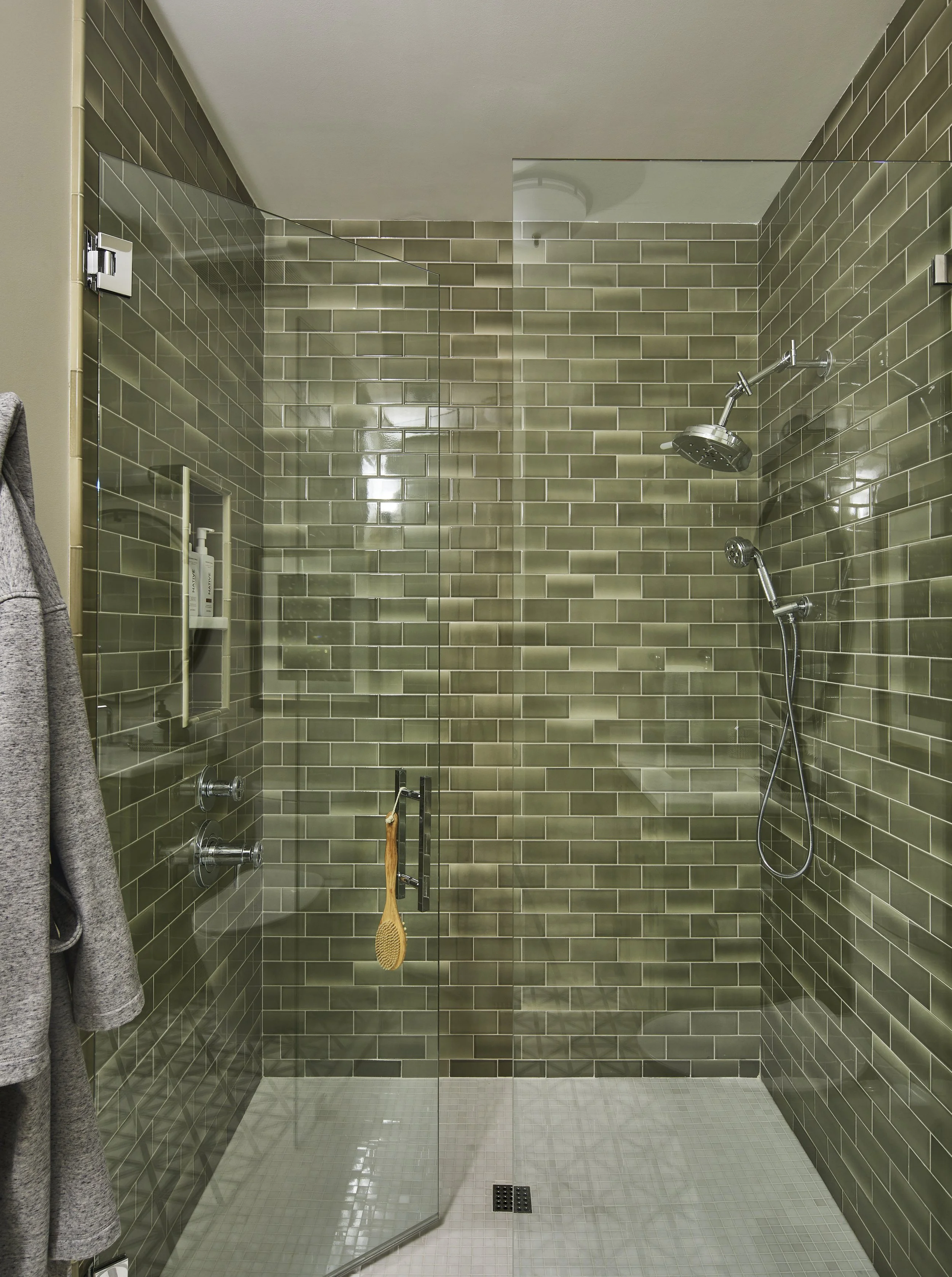 A walk-in shower with dark green subway tile walls, a glass door, a built-in shelf with toiletries, and two showerheads, one fixed and one handheld.
