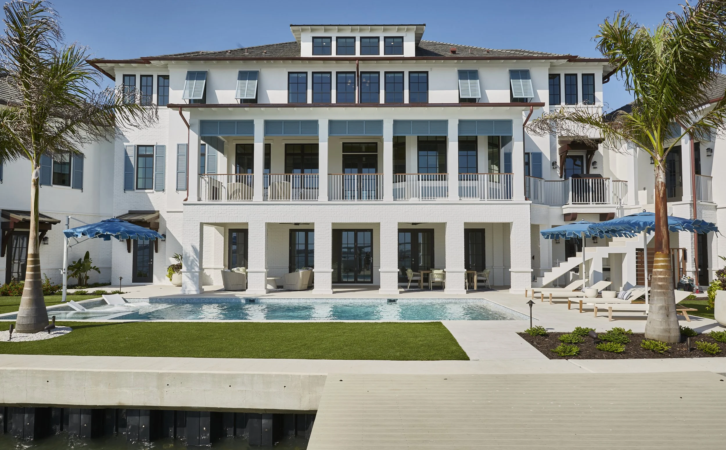 Luxury white multi-story house with pool, chairs, umbrellas, palm trees, and outdoor seating area.