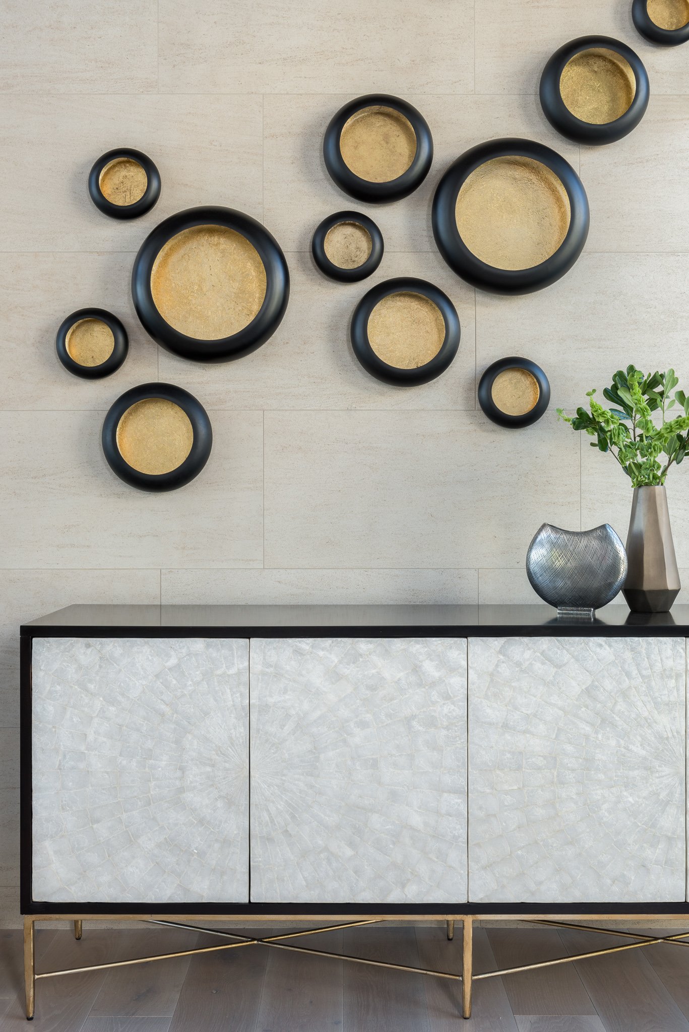 Wall decor of black circular frames with gold accent backgrounds above a modern sideboard with a textured white front and gold legs, decorated with a metallic bowl and a tall vase with green plant.