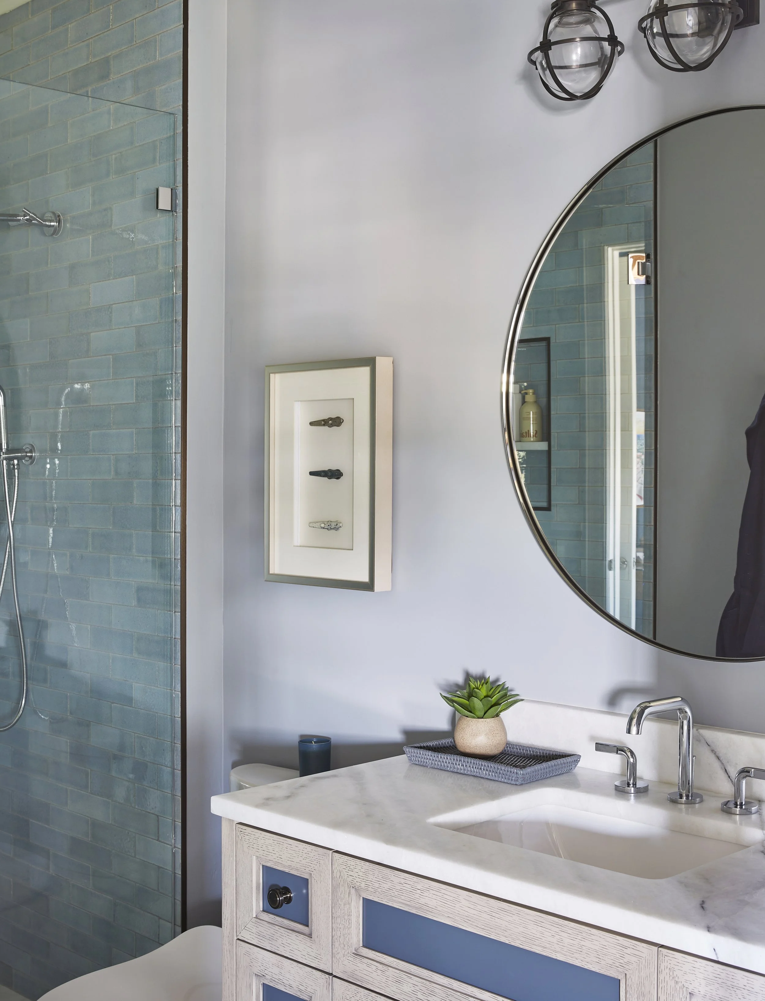 Modern bathroom interior with a white vanity, marble countertop, round mirror, and blue tile shower. Decorations include a small plant, a tray, and a framed display of keys on the wall.