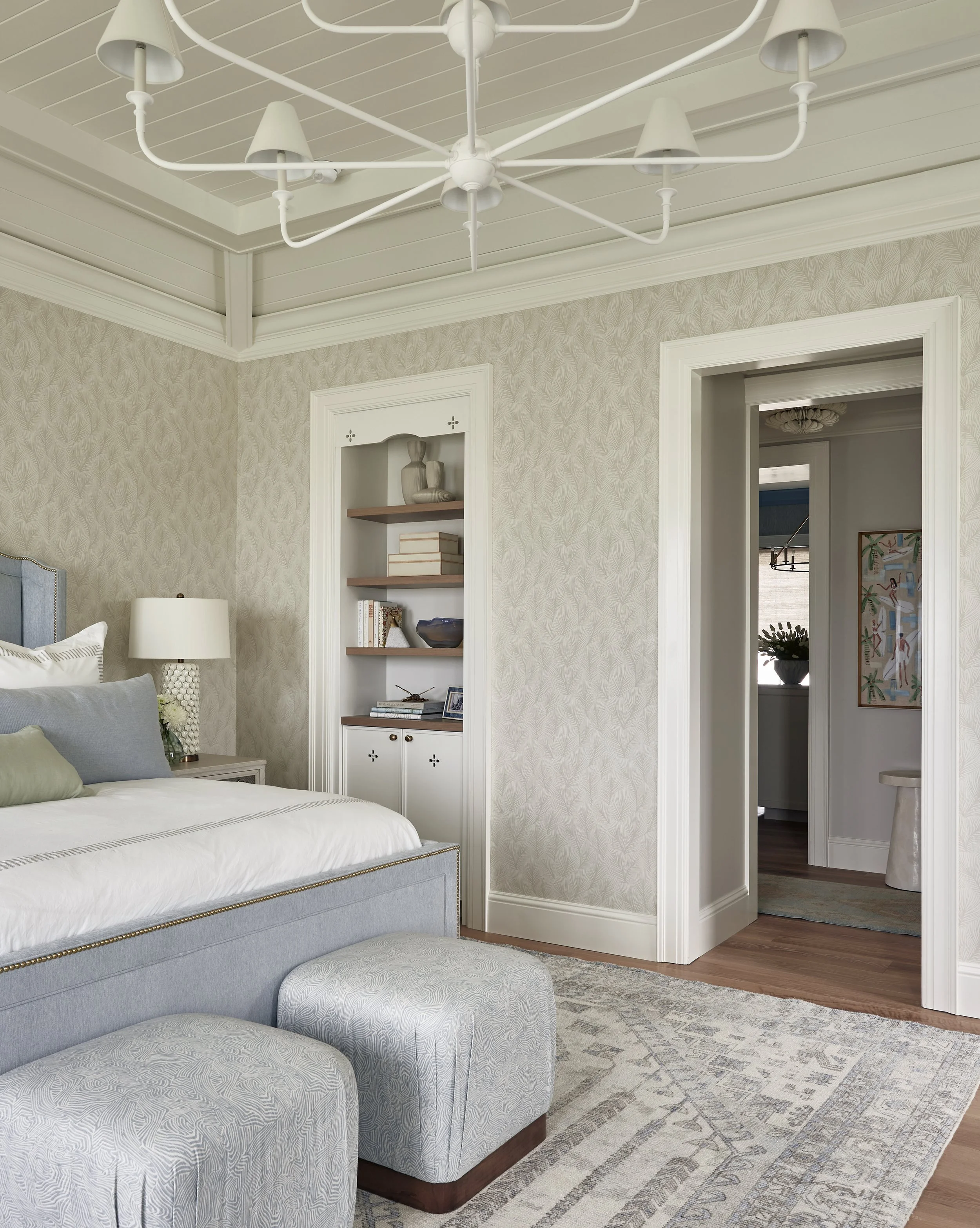 A bedroom with a light blue upholstered bed, a white nightstand, a white table lamp, and a built-in bookshelf with decorative objects and books, with an inlaid wood floor and a patterned area rug.