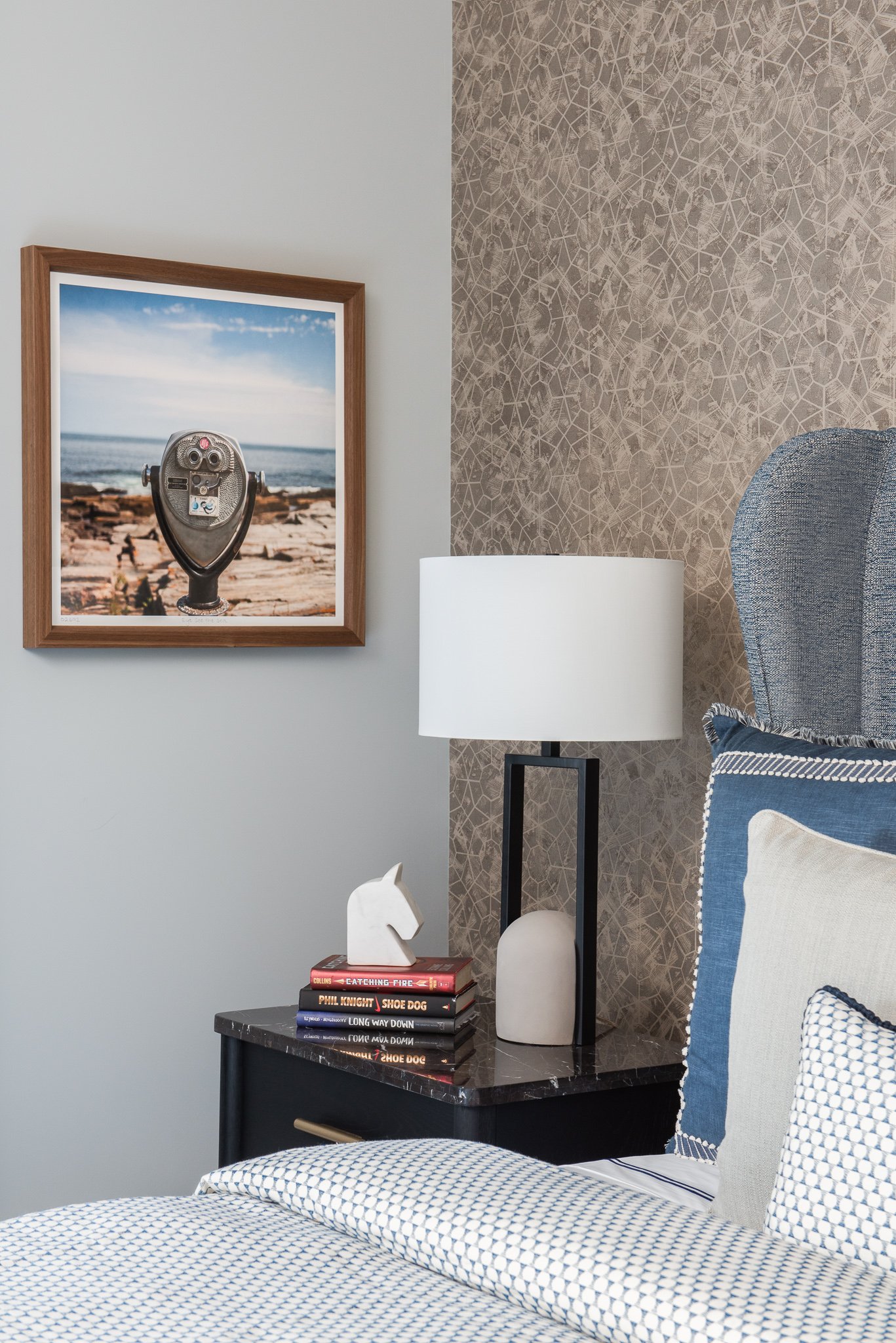 A bedroom with a bed, a navy blue pillow with beige trim, a dark nightstand, a white horse figurine, a stack of books, a modern lamp, and a framed picture of a coin-operated viewing scope on a rocky beach with ocean in the background.