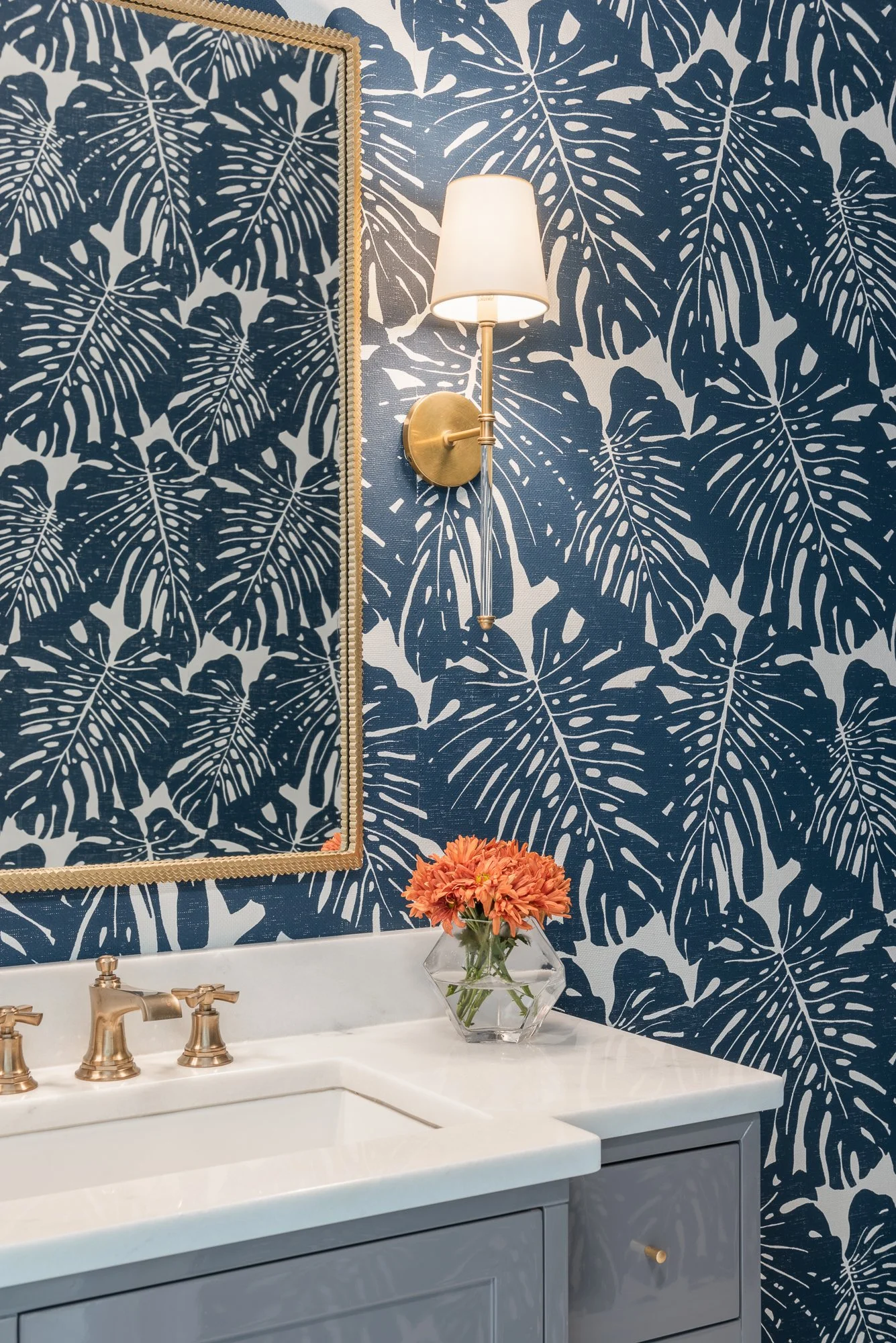 Bathroom with navy blue tropical leaf wallpaper, gold lighting, a mirror, a white sink with gold fixtures, and a glass vase with orange flowers on the countertop.