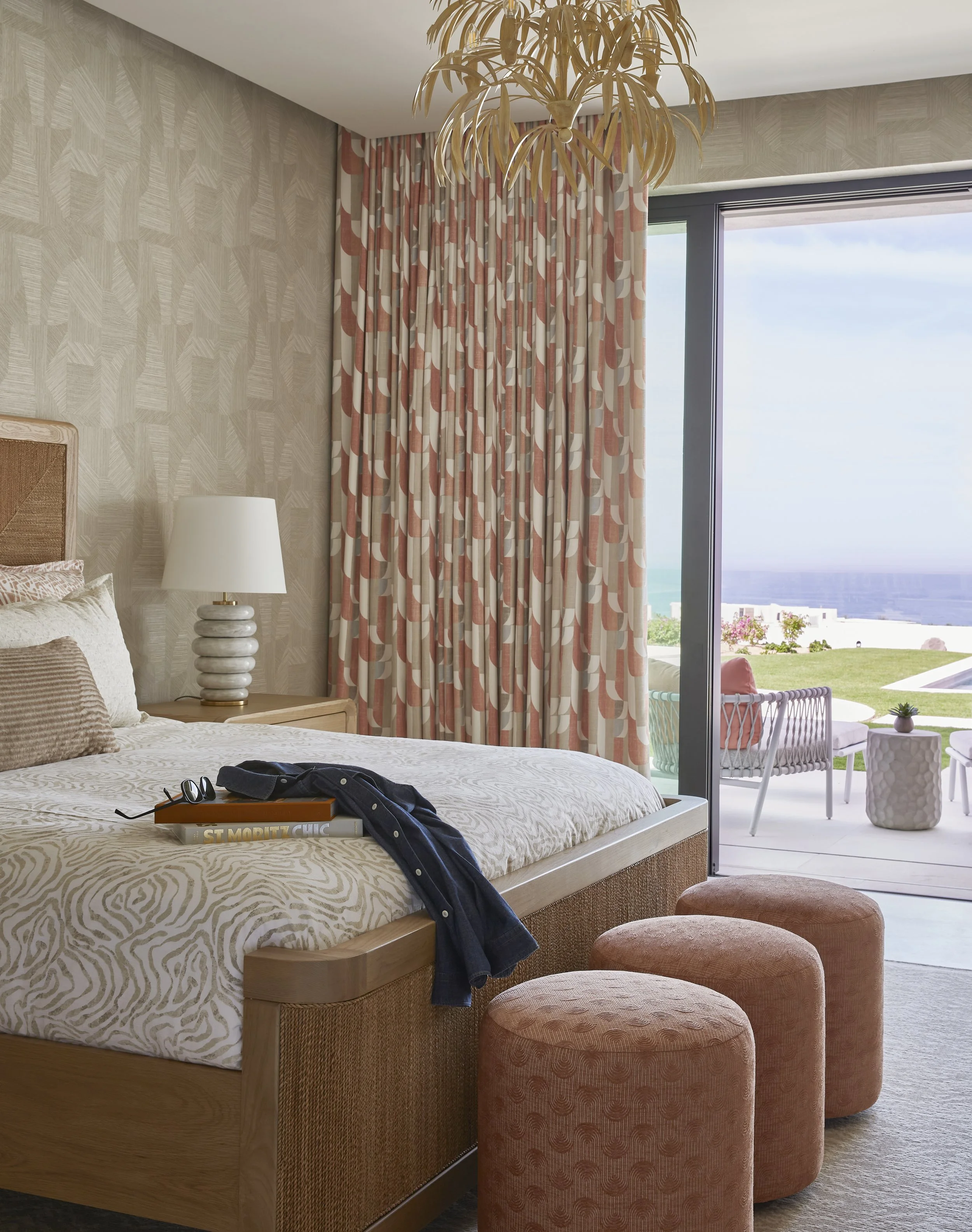 A bedroom with a large window showing a balcony with outdoor furniture and a garden view. The bed has a wooden frame, and there are three pink upholstered ottomans at the foot of the bed. The walls have textured wallpaper, and there is a lamp with a 