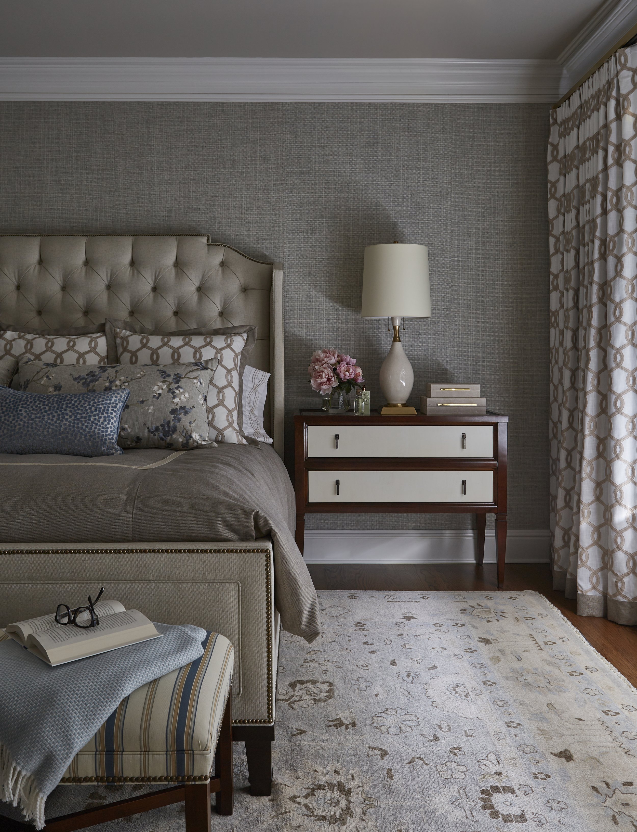 A bedroom with a beige upholstered bed, decorative pillows, a wooden nightstand with a white lamp, pink flowers, books, and a vase, a patterned rug, and cream-colored curtains with a geometric pattern.