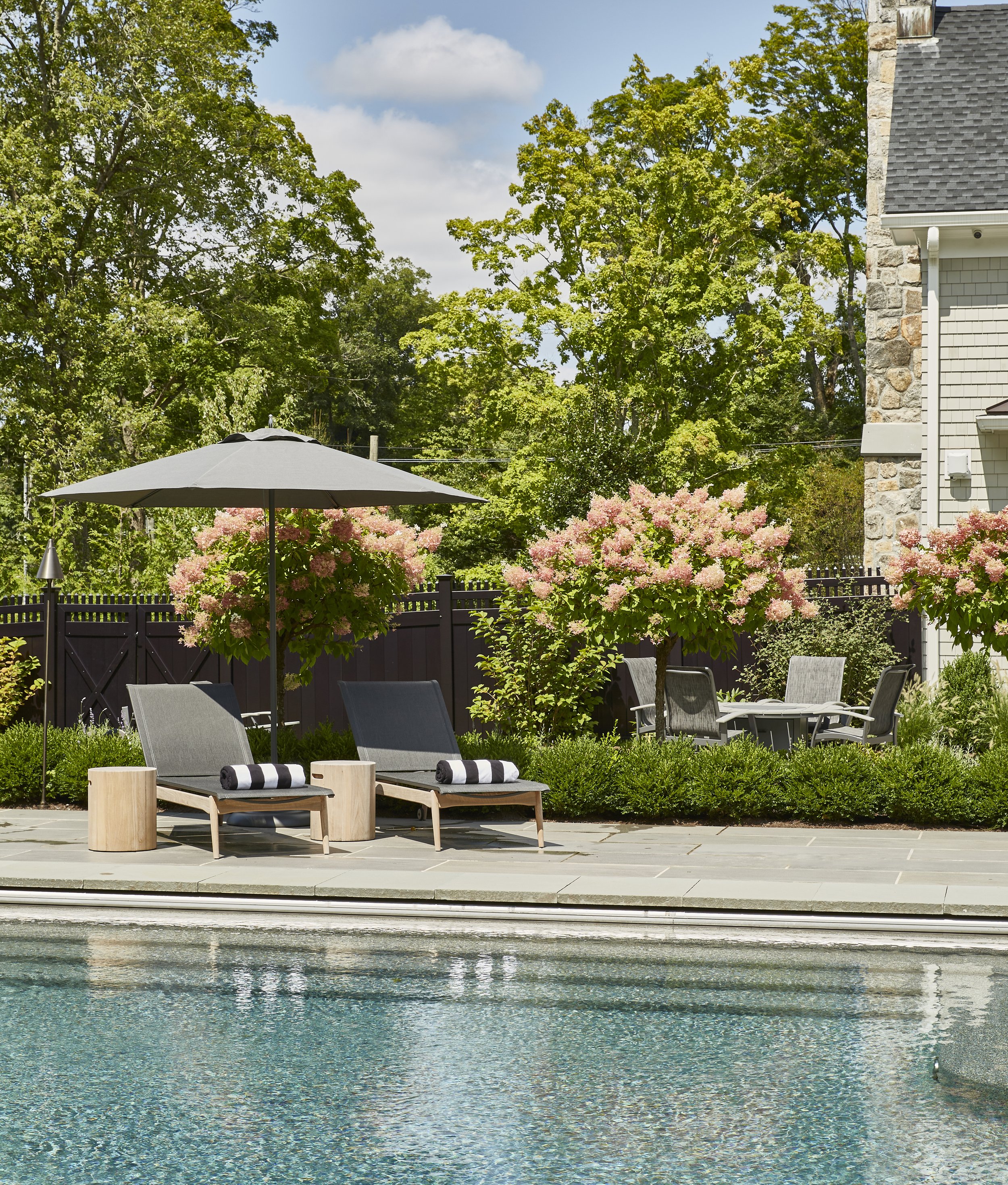 A backyard with a swimming pool, two lounge chairs with rolled towels under a large gray patio umbrella, and a garden with pink flowering bushes and green trees in the background.