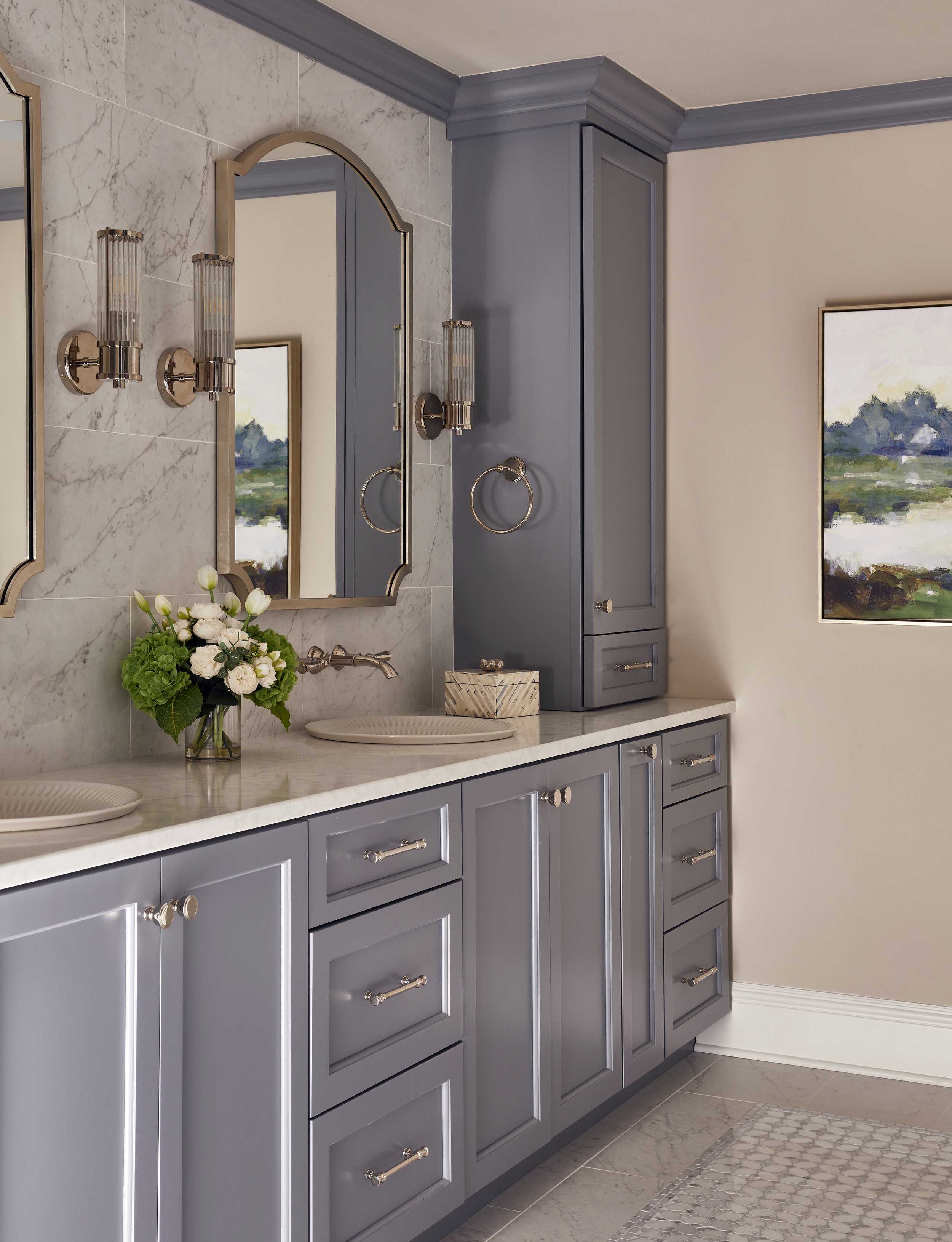 Bathroom with blue cabinets, marble countertop, two oval mirrors, wall-mounted sconces, and a painting of a landscape on the wall.