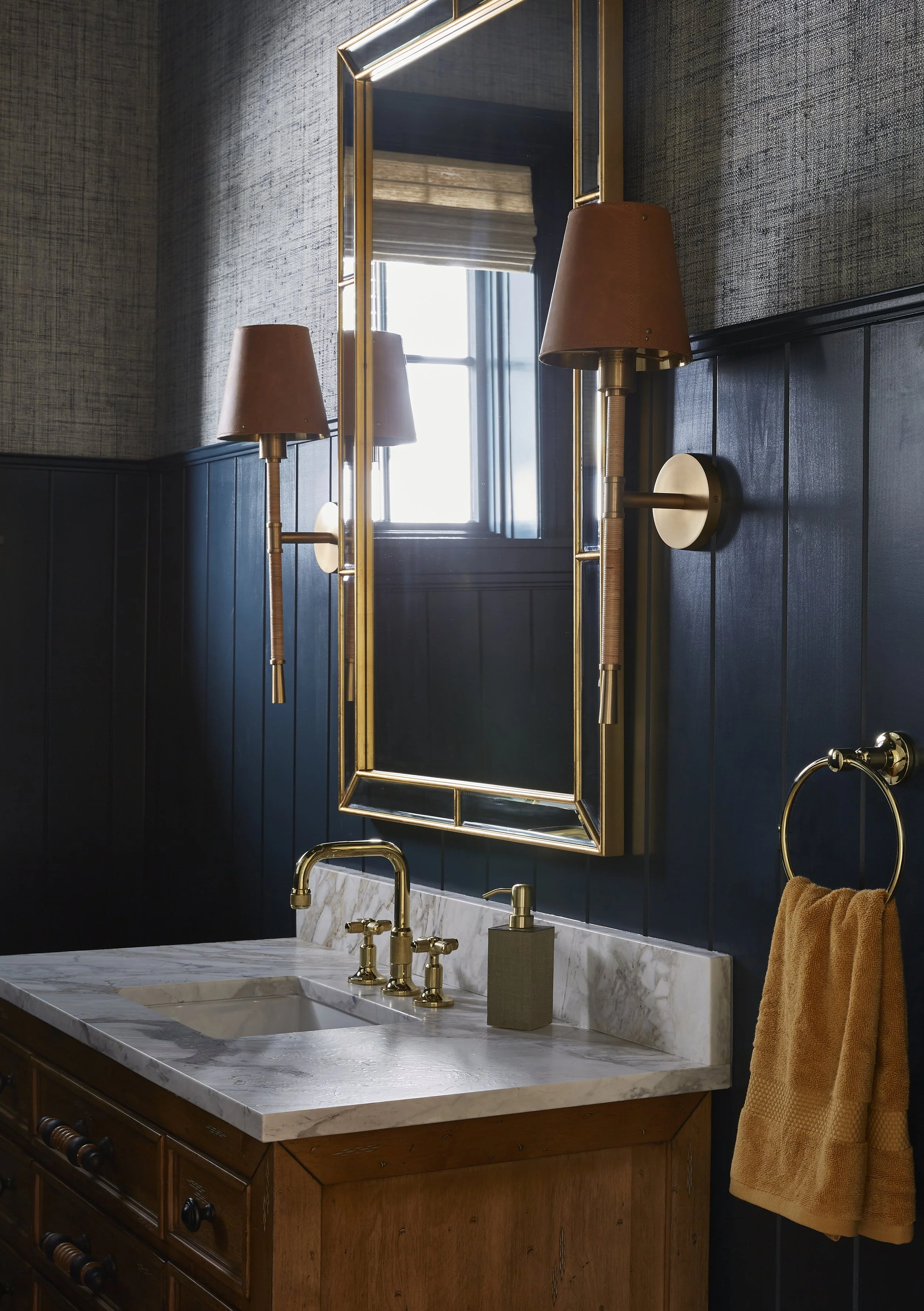 A bathroom sink with a marble countertop, gold fixtures, a rectangular mirror with a gold frame, wall-mounted lamps, a window with a wooden shade, and an orange towel hanging on a gold towel ring.