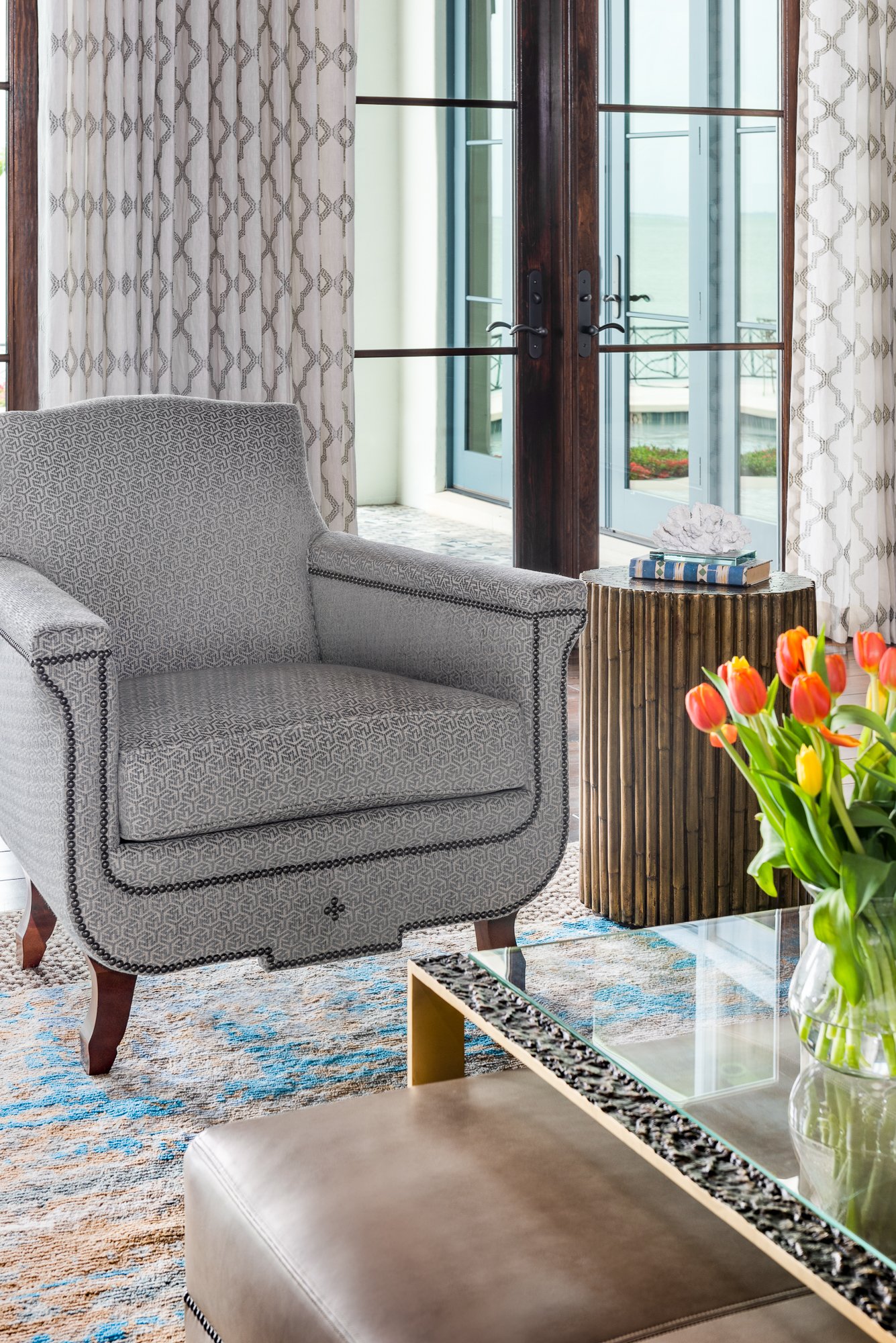A living room with a patterned armchair, a side table with books and a coral decoration, a glass-top coffee table with a vase of orange and yellow tulips, and a large glass door with curtains leading to a balcony or outdoor area.