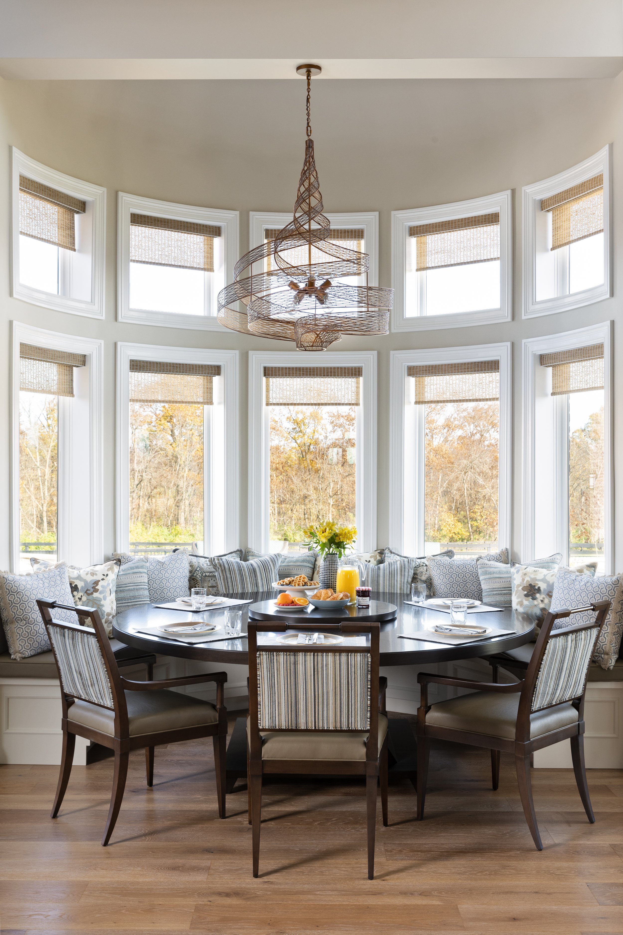 A cozy dining area with a round table set for breakfast, surrounded by chairs and a built-in window seat with pillows, near large windows with wood blinds, and a modern pendant light overhead.