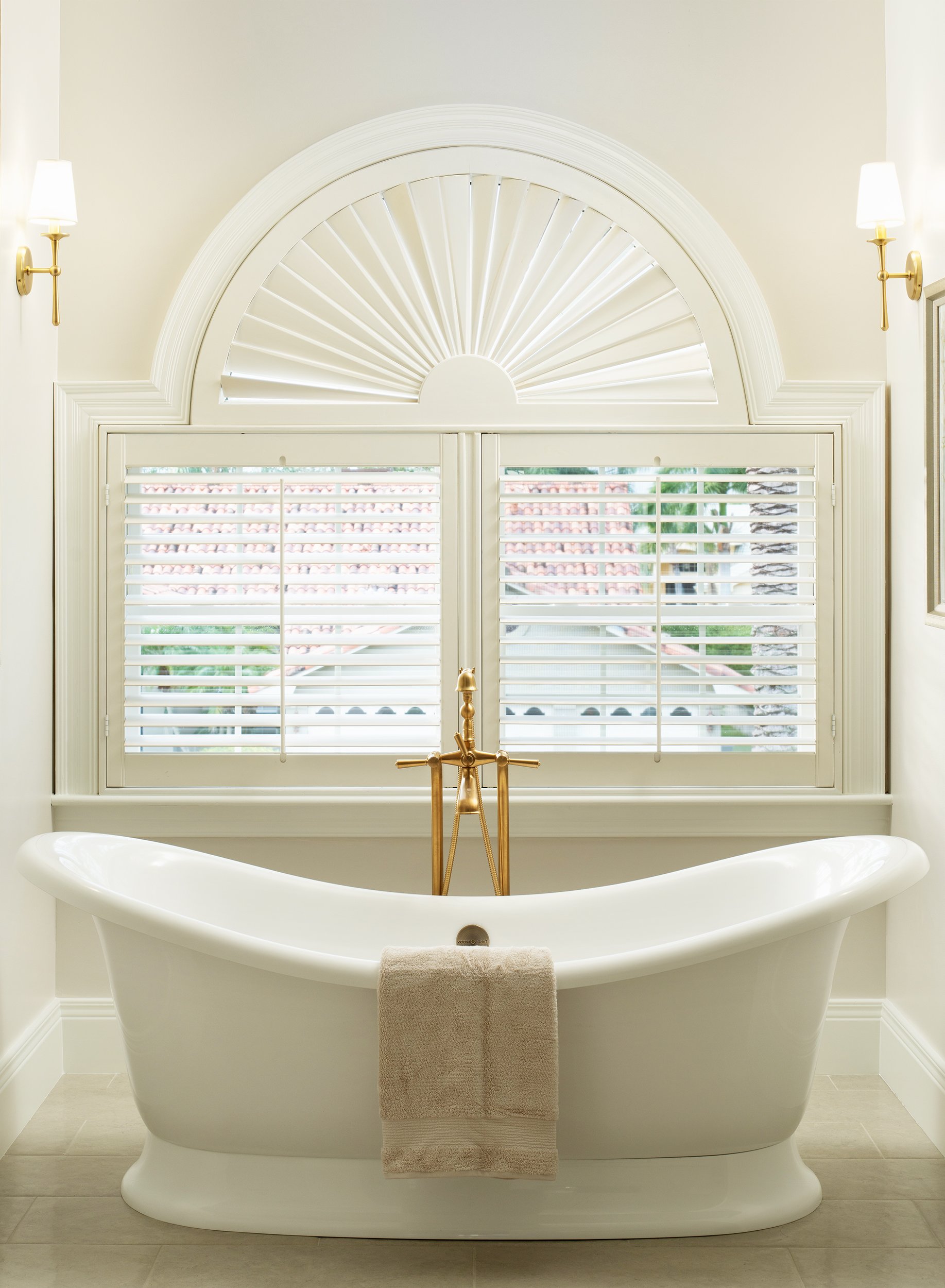 Elegant bathroom with a white clawfoot tub, a beige towel, a window with white shutters, and wall-mounted lights.