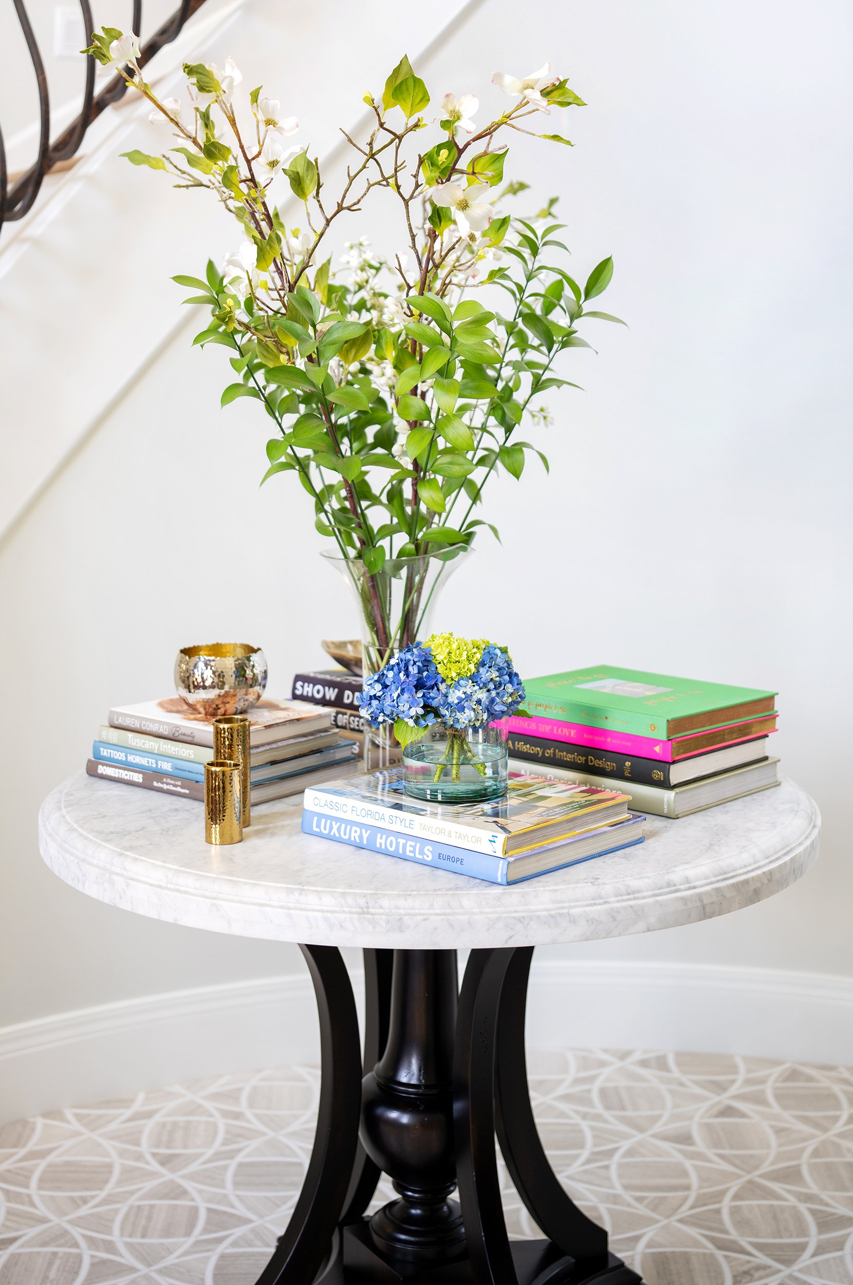 A round marble-top table with a black pedestal base, topped with two clear vases of flowers, stacks of books, and small decorative objects, set against a neutral background.