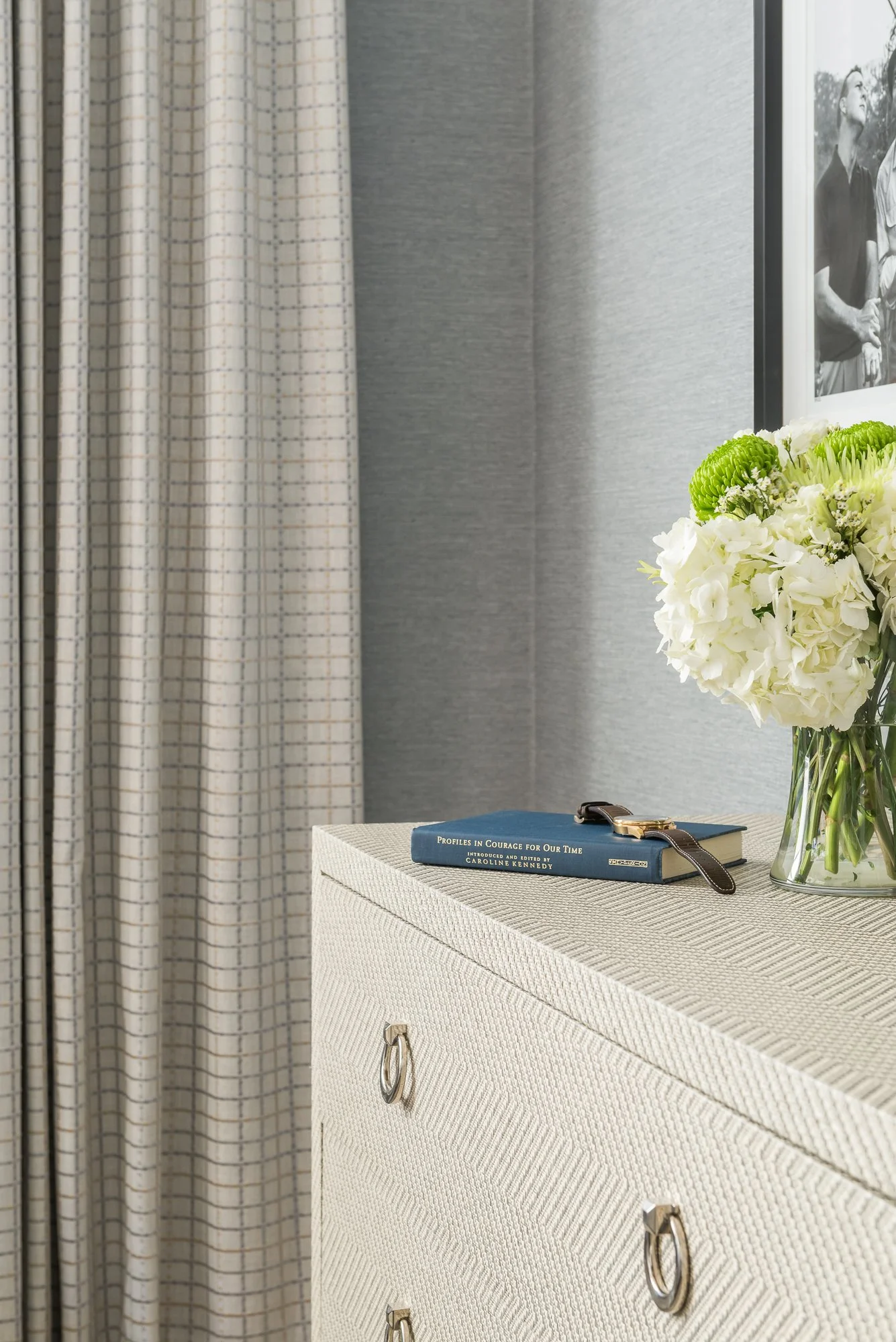 Close-up of a cream-colored textured dresser with silver ring pulls, a book titled 'Profiles in Courage For Our Time' with a watch on top, and a glass vase with white and green flowers, against a gray wall, partial black-and-white photograph in the b