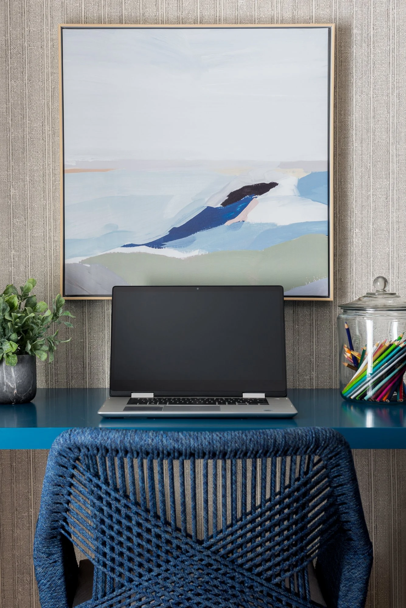 A home office setup featuring a blue desk, a wired laptop with a blank screen, a blue woven chair, a potted plant, a glass container filled with colored pens, and a large abstract landscape painting on a patterned wallpaper wall.
