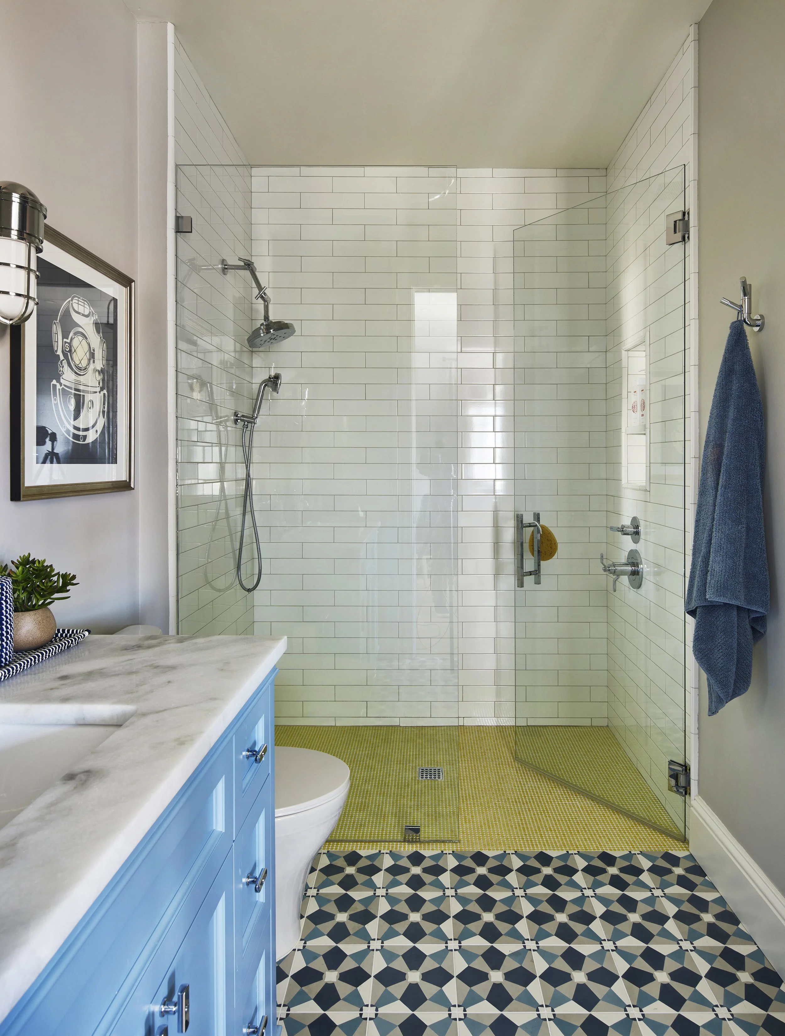 Bathroom with a glass-enclosed shower, white subway tile walls, a marble countertop vanity with blue cabinets, patterned floor tiles, and a blue towel hanging on the wall.