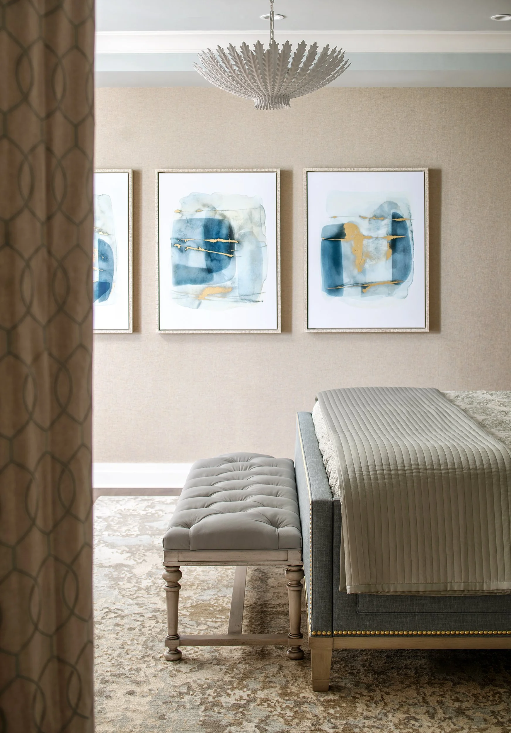A room with a bed, nightstand, three abstract blue and gold paintings on the wall, a decorative chandelier, and a patterned rug.