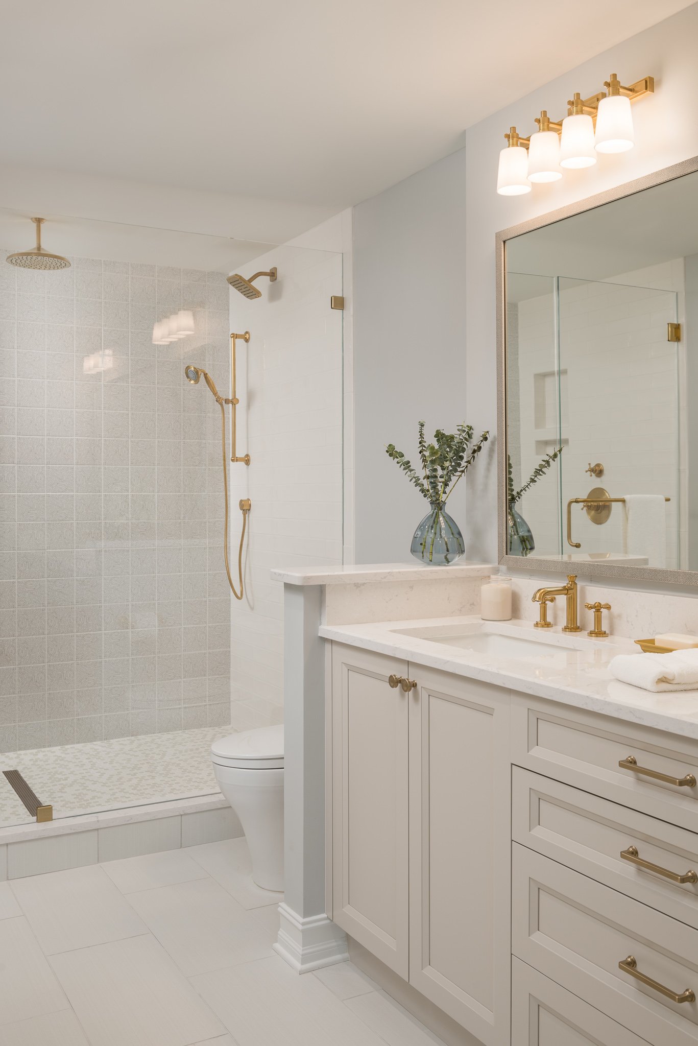 Modern bathroom with a walk-in shower, white cabinetry, a large mirror, and gold fixtures, including a faucet, shower fixtures, and cabinet handles, with a vase of greenery on the counter.