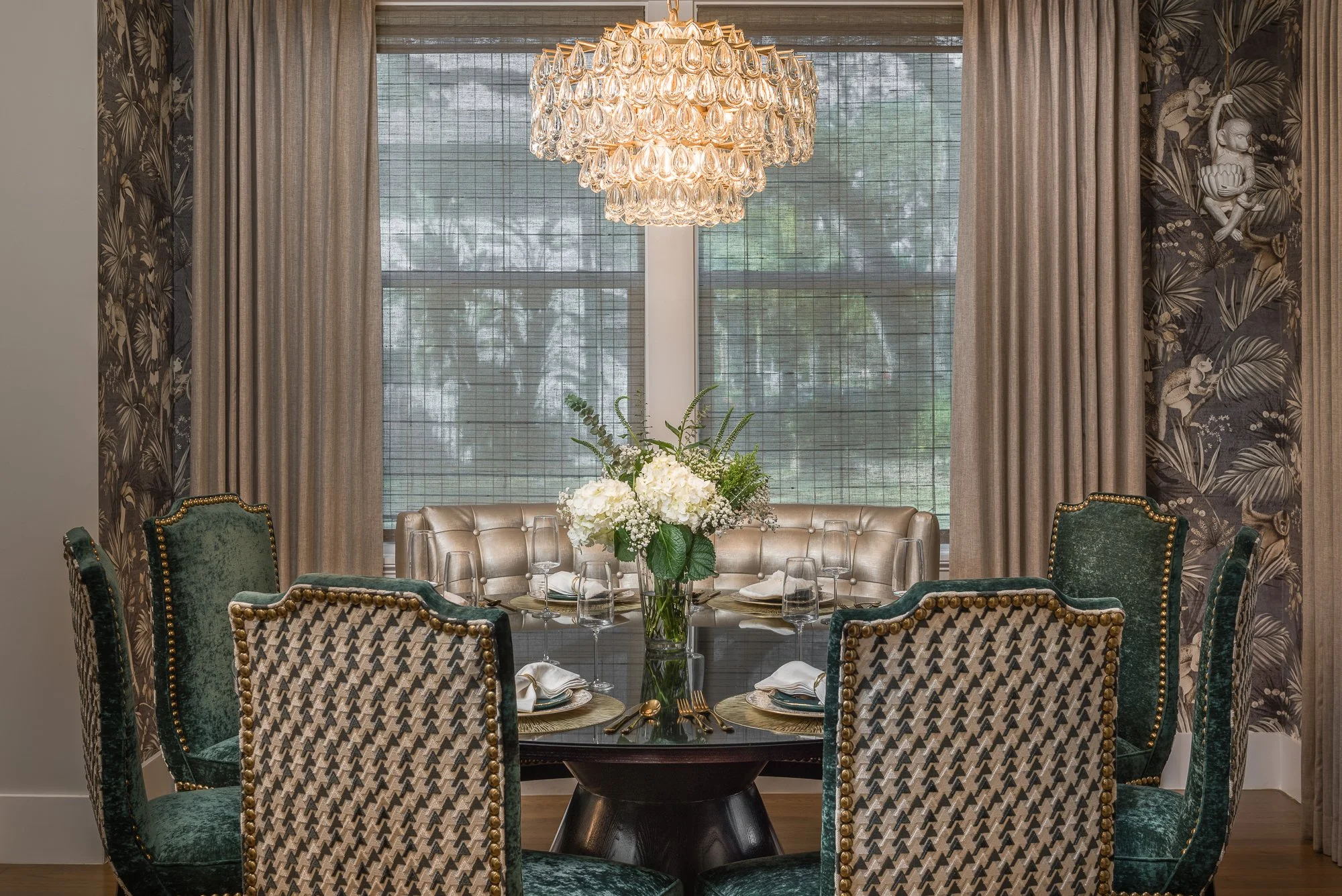 Elegant dining room with a round table, six upholstered chairs, a large floral centerpiece, a chandelier, and windows with curtains.