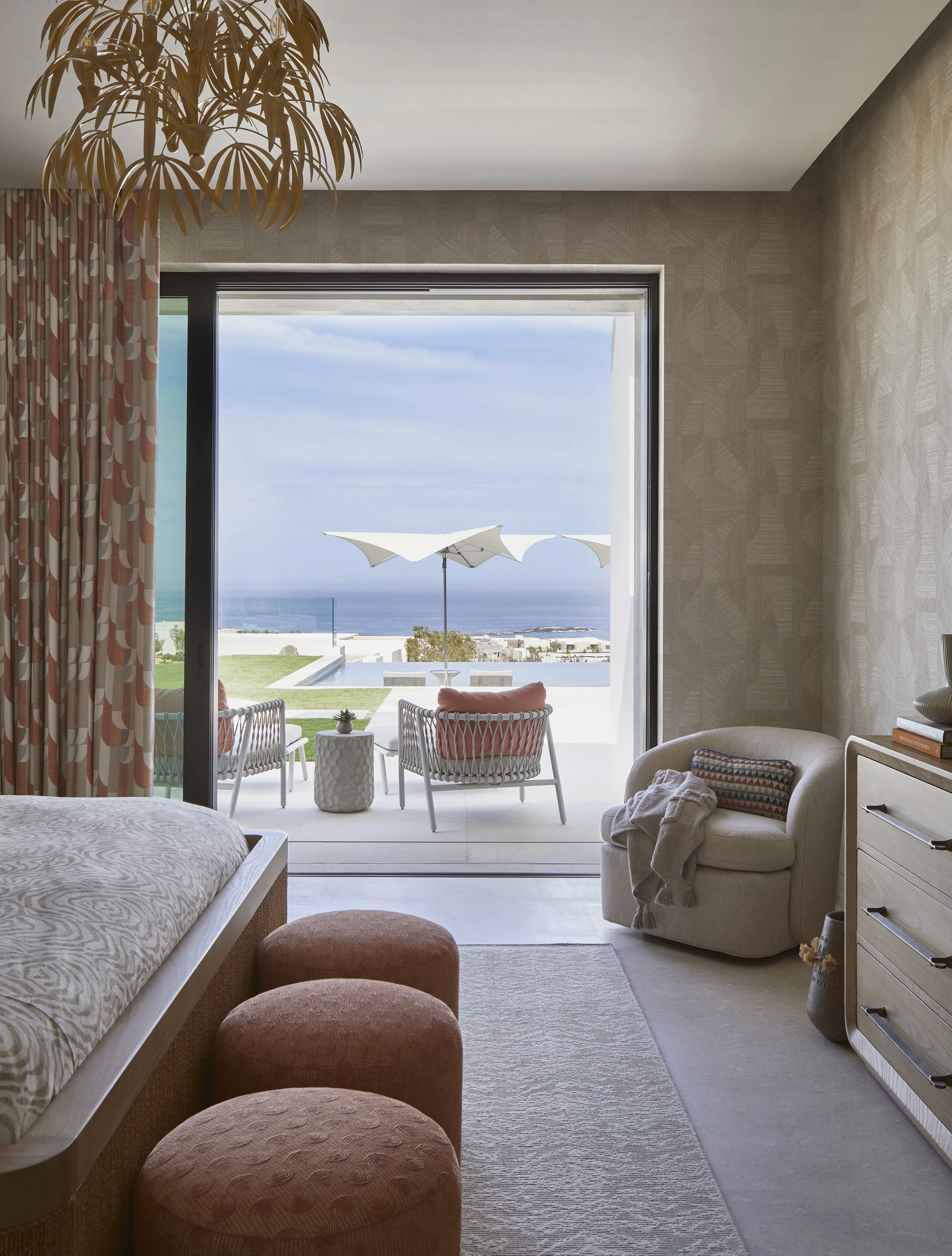 A bedroom with a large sliding glass door opening to a balcony overlooking the ocean, with outdoor furniture including chairs, a small table, and umbrellas.