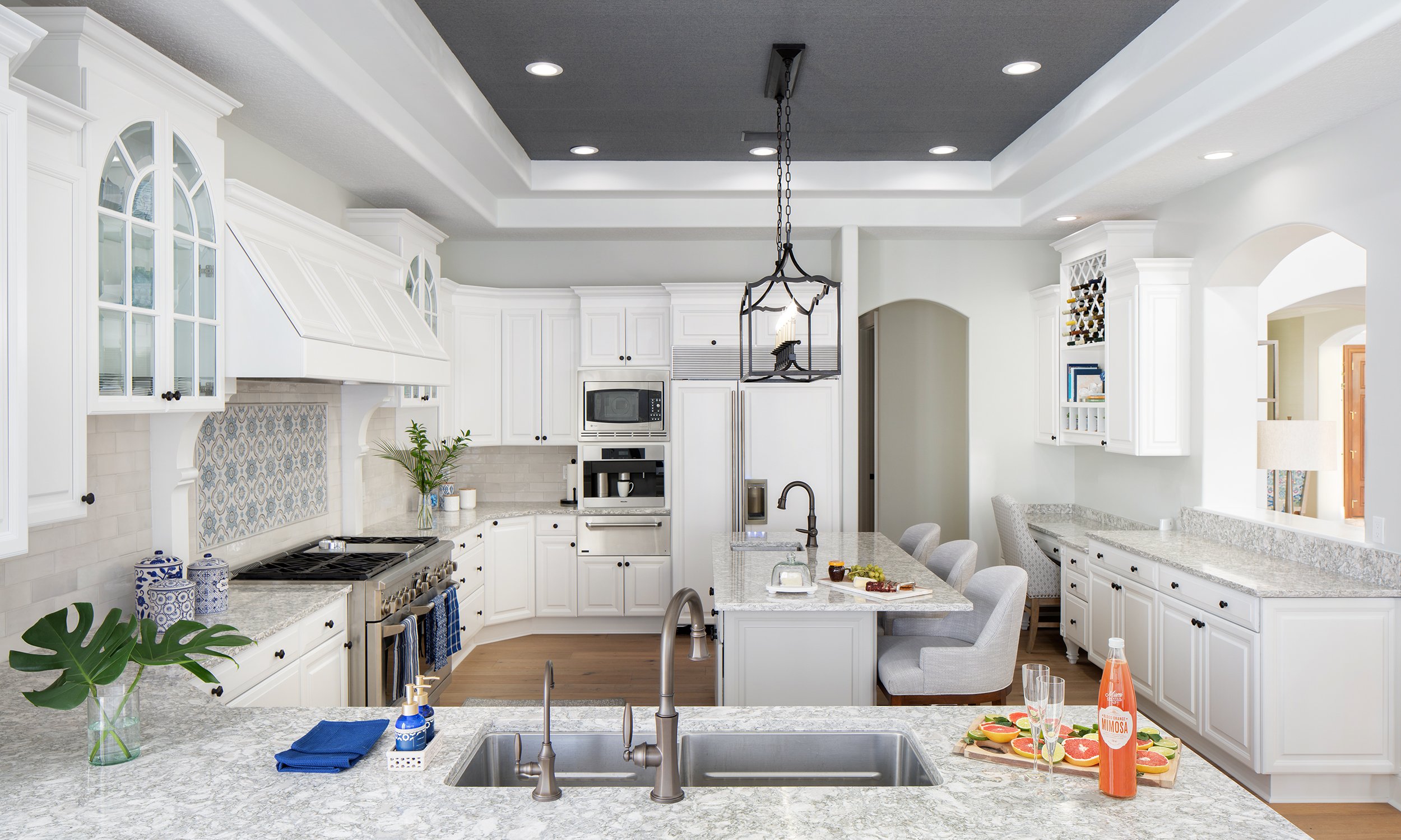Modern white kitchen with granite countertops, stainless steel appliances, an island with bar seating, and decorative plants and accessories.