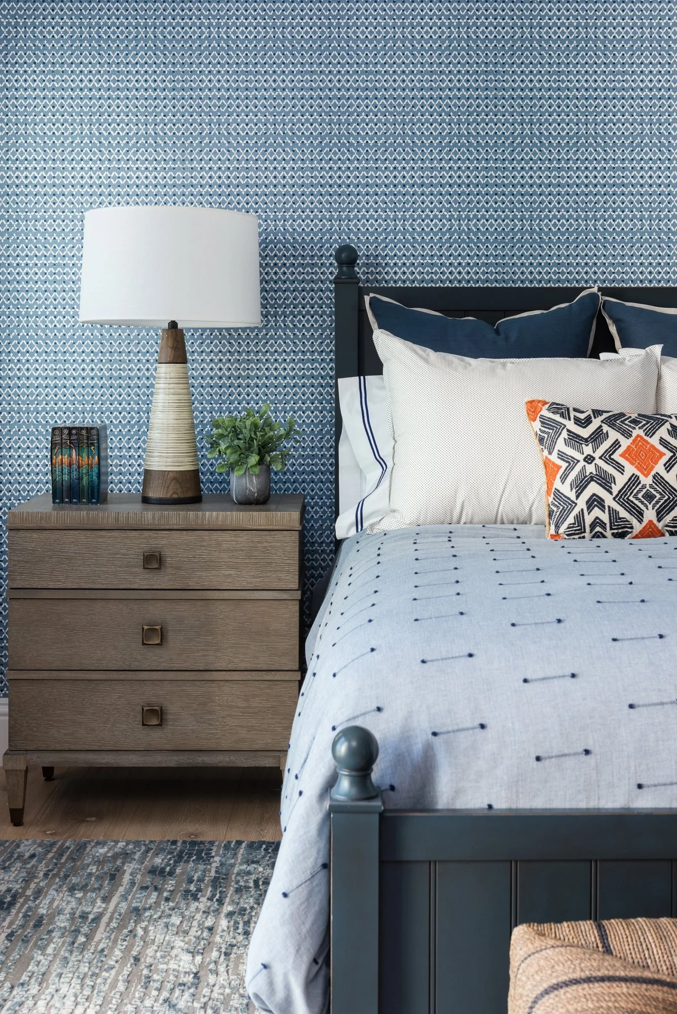 Bedroom with blue patterned wallpaper, a black bed frame, a bedside table with a lamp, a plant, and a set of books. The bed has white and navy bedding with decorative pillows.