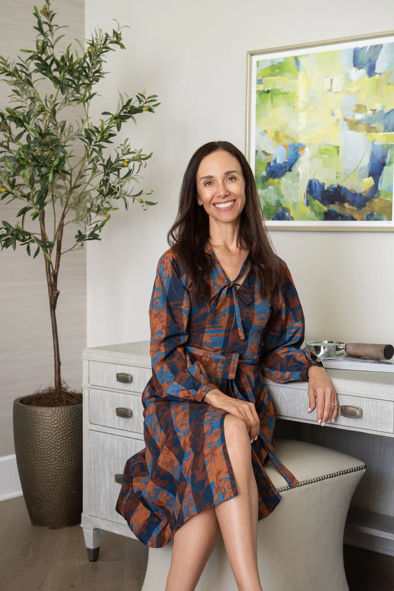 A woman with long dark hair smiling, sitting on a white stool in front of a desk, wearing a floral dress with blue and brown colors. There is a large potted plant and colorful abstract painting behind her.