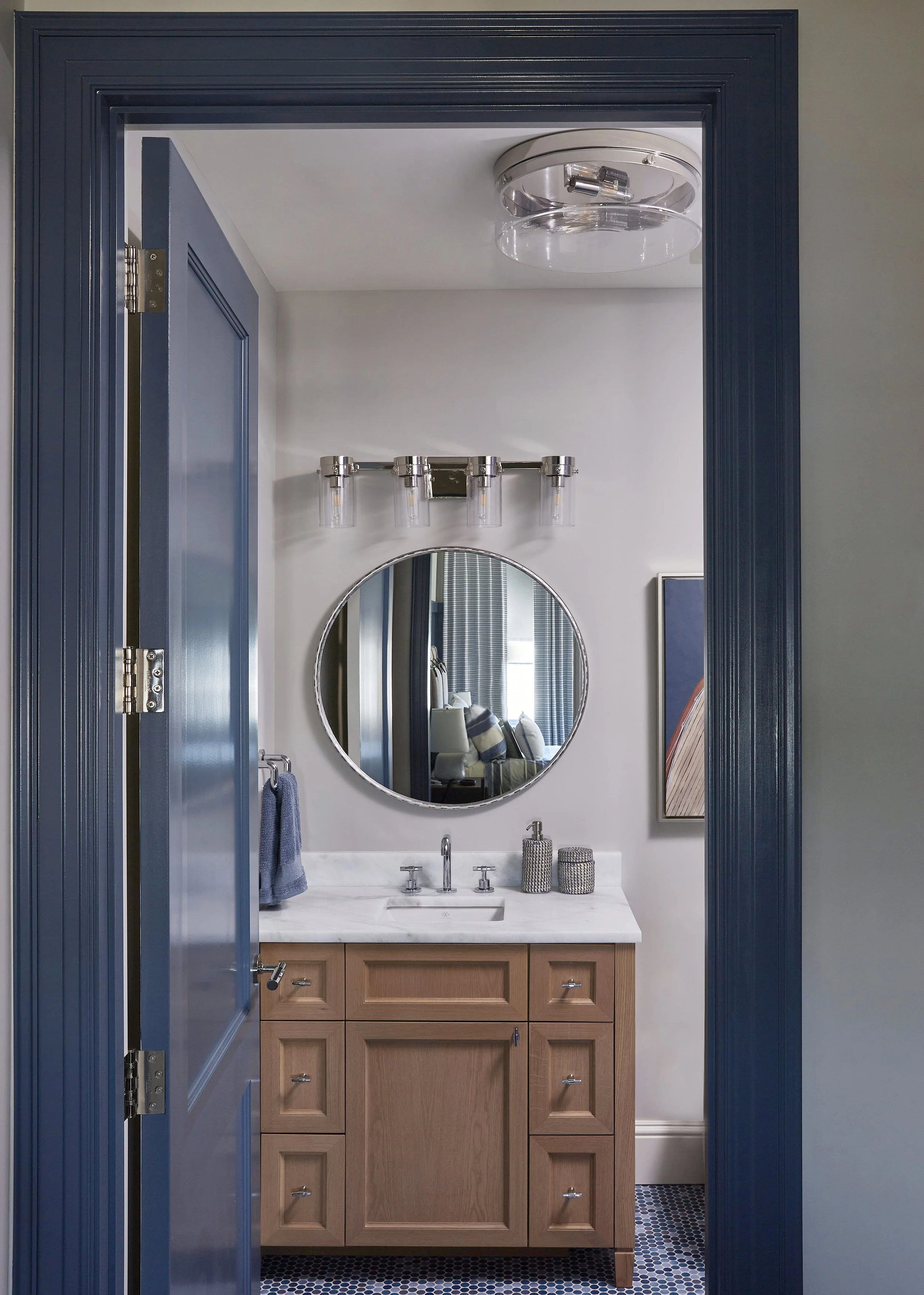 Bathroom vanity with a round mirror, wooden cabinet, marble countertop, and modern light fixture, viewed through a blue door frame.