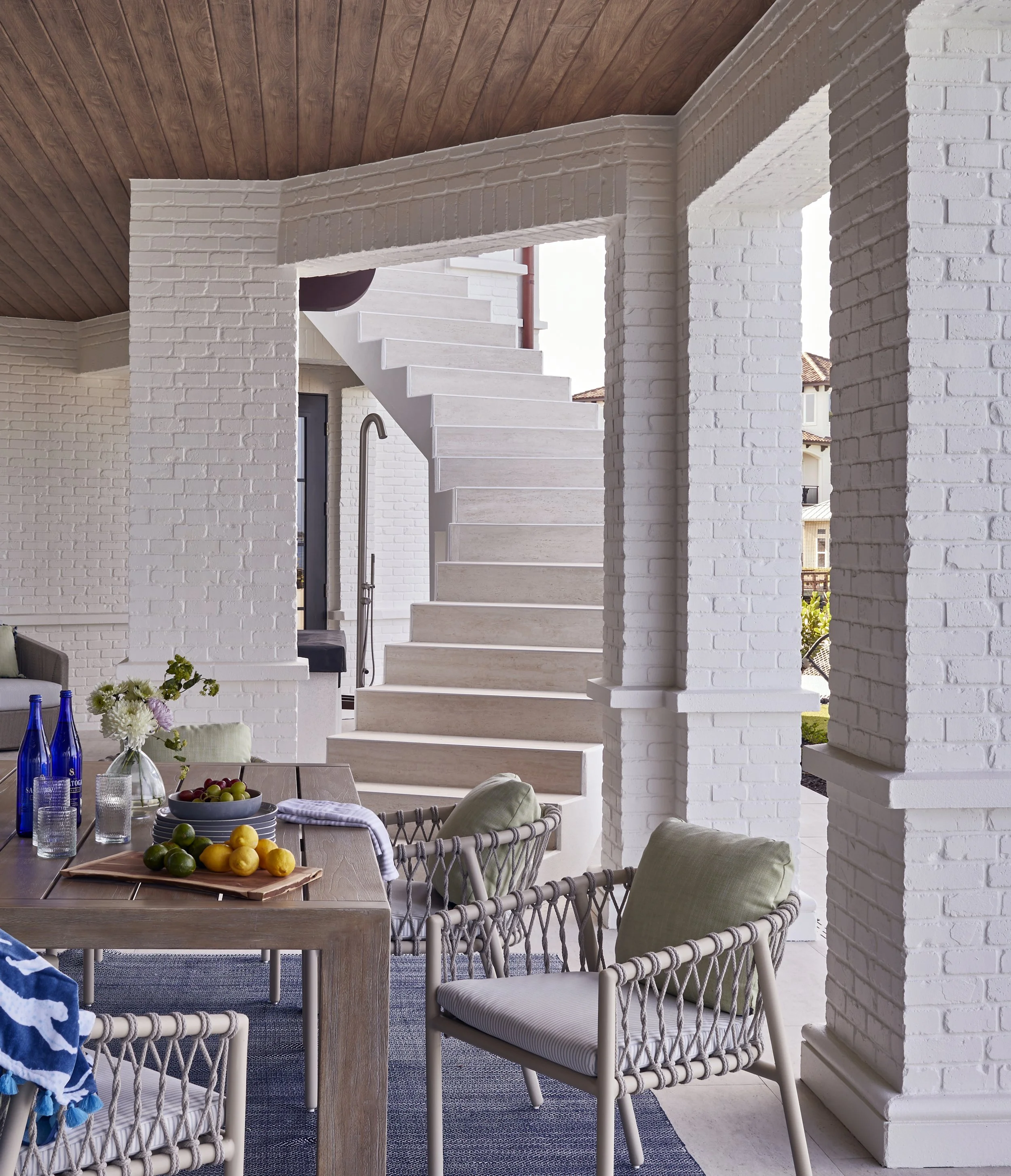 Interior view of a modern outdoor living space with white brick walls, a wooden ceiling, and a staircase leading upstairs. There is a dining table with fruit and bottles, surrounded by cushioned chairs with green and neutral pillows.