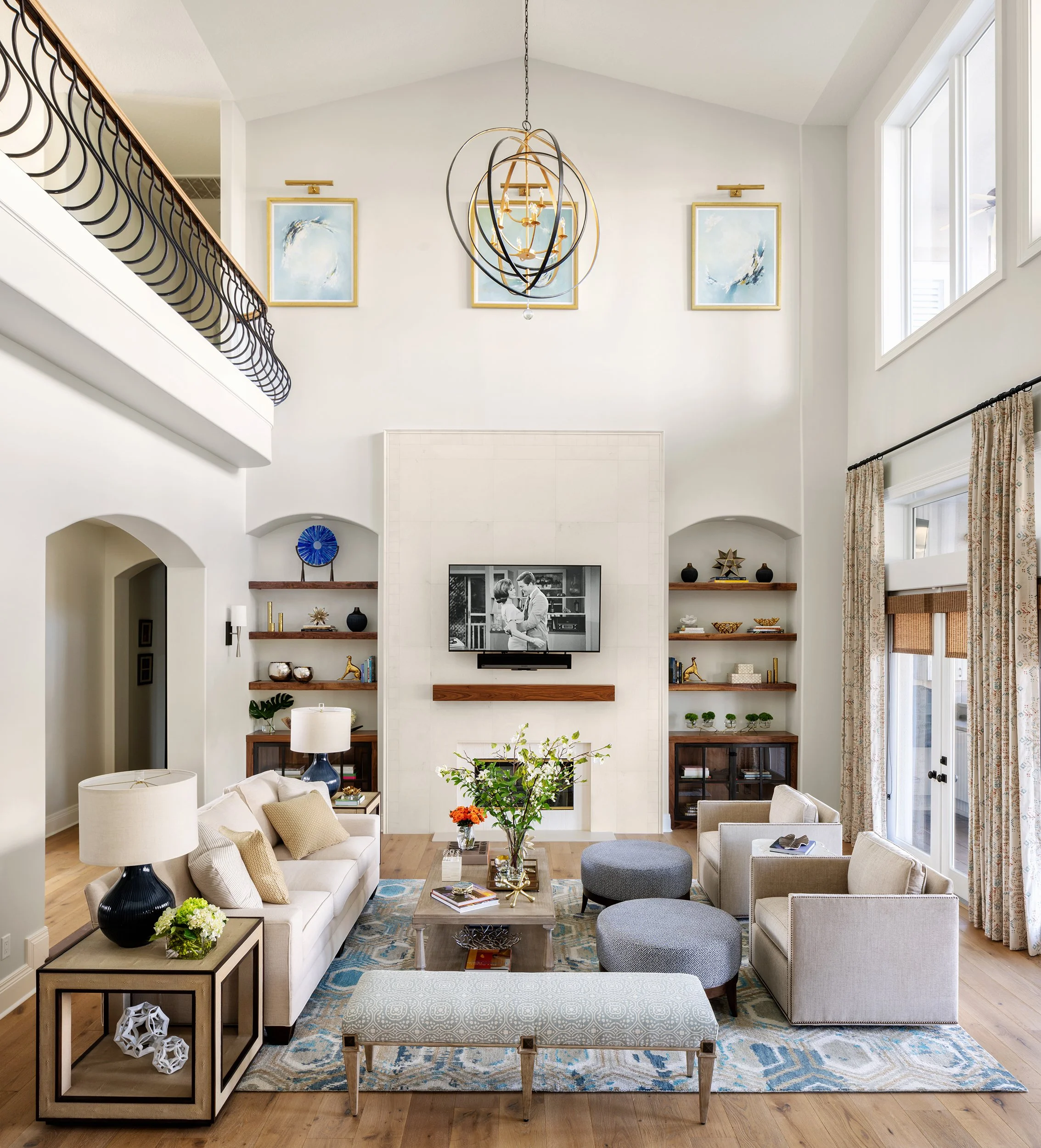 A spacious living room with a high vaulted ceiling, a large flat-screen TV above a fireplace, and built-in wall shelves on either side. The room has a modern, elegant decor with a white sofa, armchairs, ottomans, and a coffee table decorated with boo