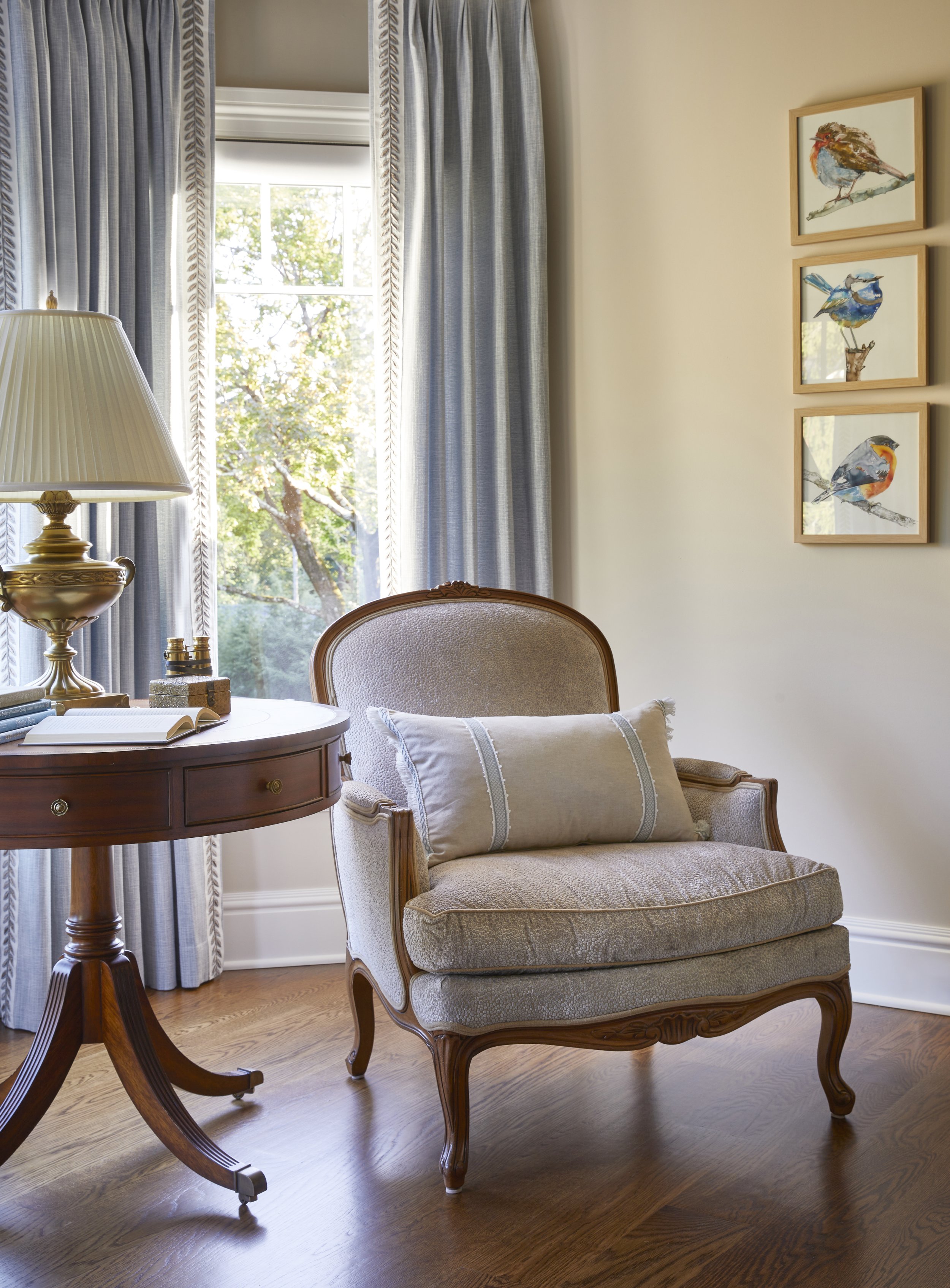 A cozy reading nook with a vintage beige armchair and pillow beside a round wooden side table with a brass lamp and books. Behind the chair, a large window with blue curtains shows green trees outside. On the wall, three framed watercolor paintings o