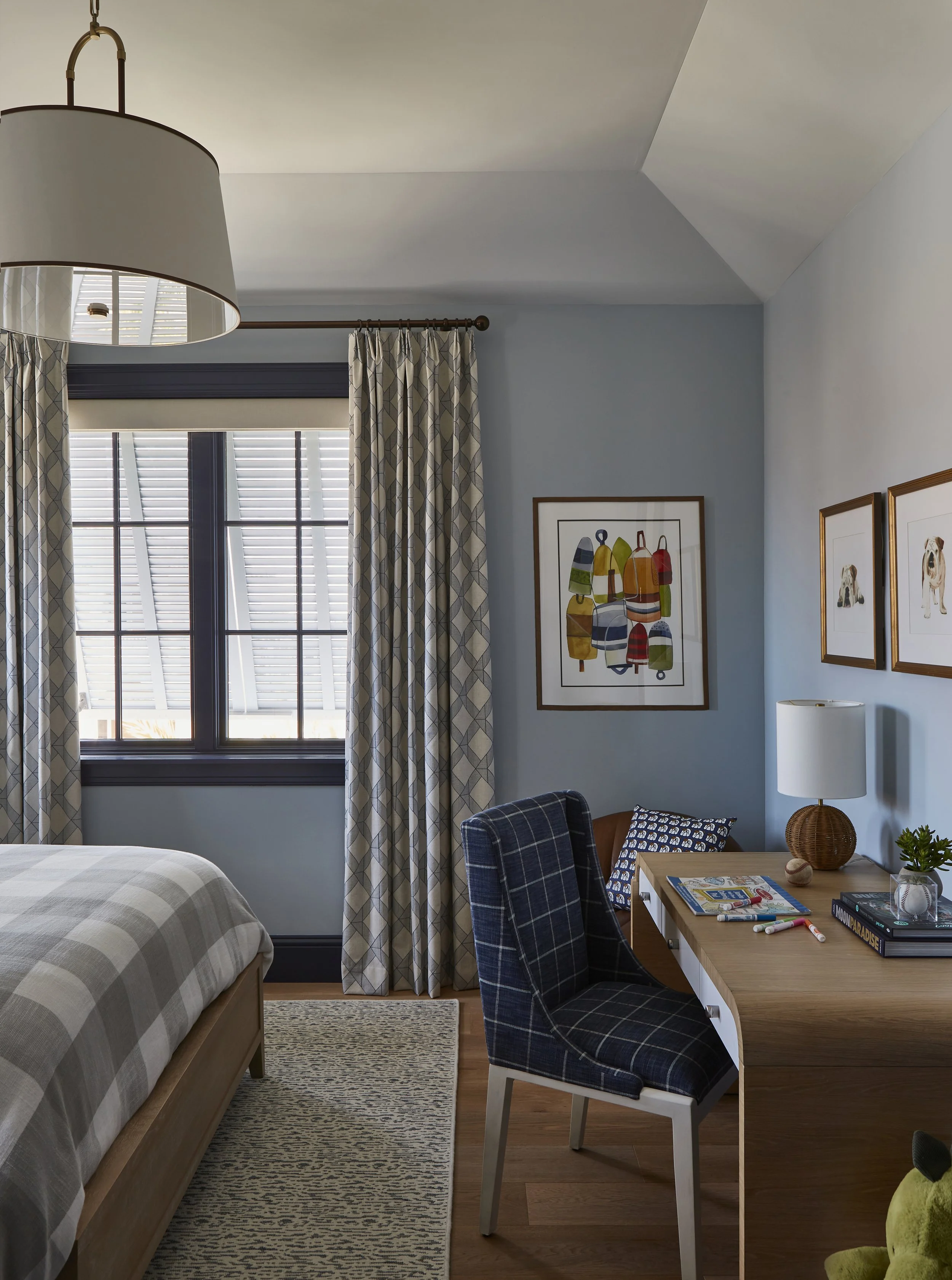 A bedroom with a large window, checkered curtains, a bed with a checkered comforter, a wooden desk with a bookshelf, a black plaid chair, framed artwork on the walls, a white lamp with a woven base, and various books and decorative items on the desk.