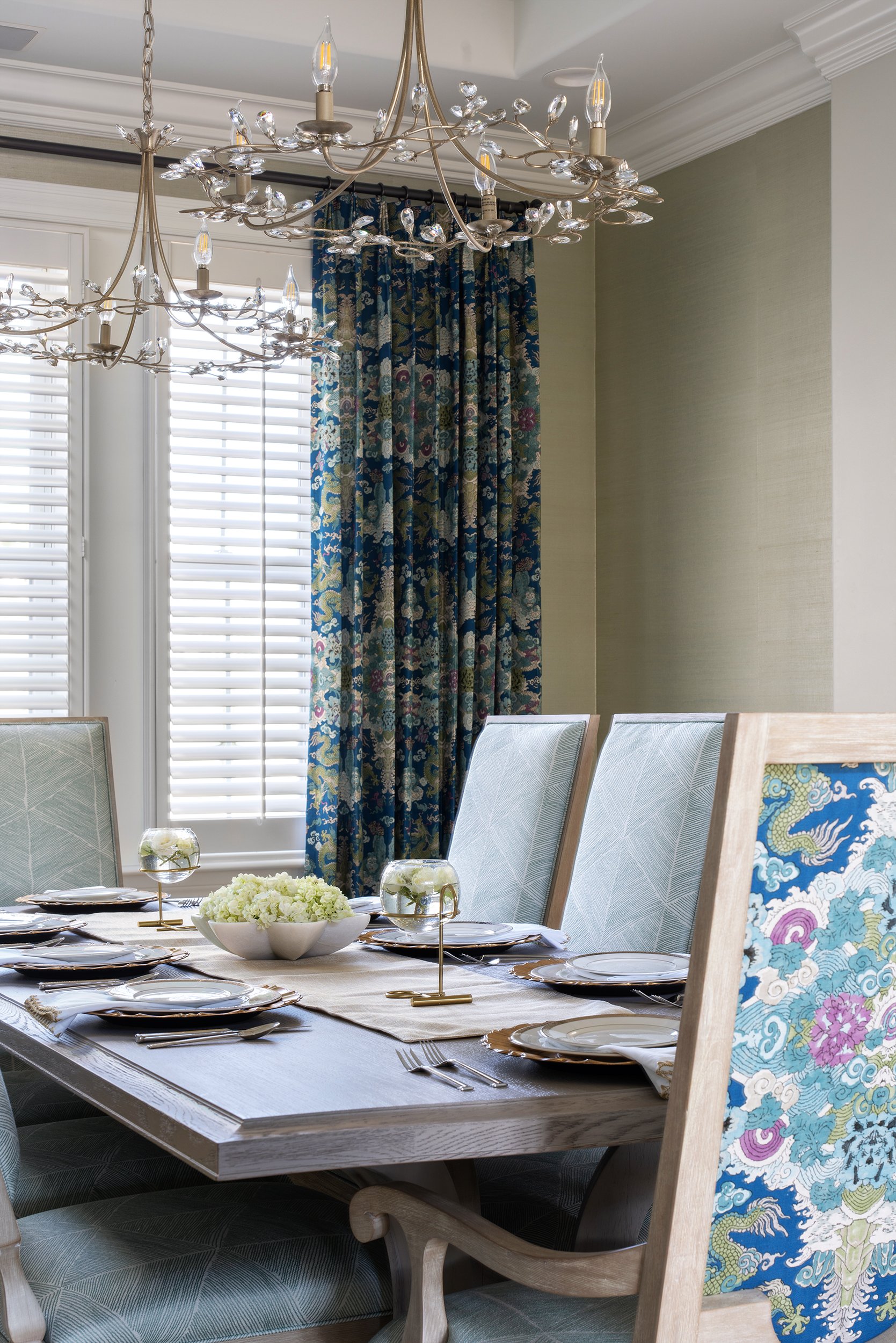 Elegant dining room with a wooden table set with white plates, silverware, and glass centerpieces. There are upholstered chairs with patterned fabric and a large window with white shutters and blue floral curtains. A decorative chandelier hangs above