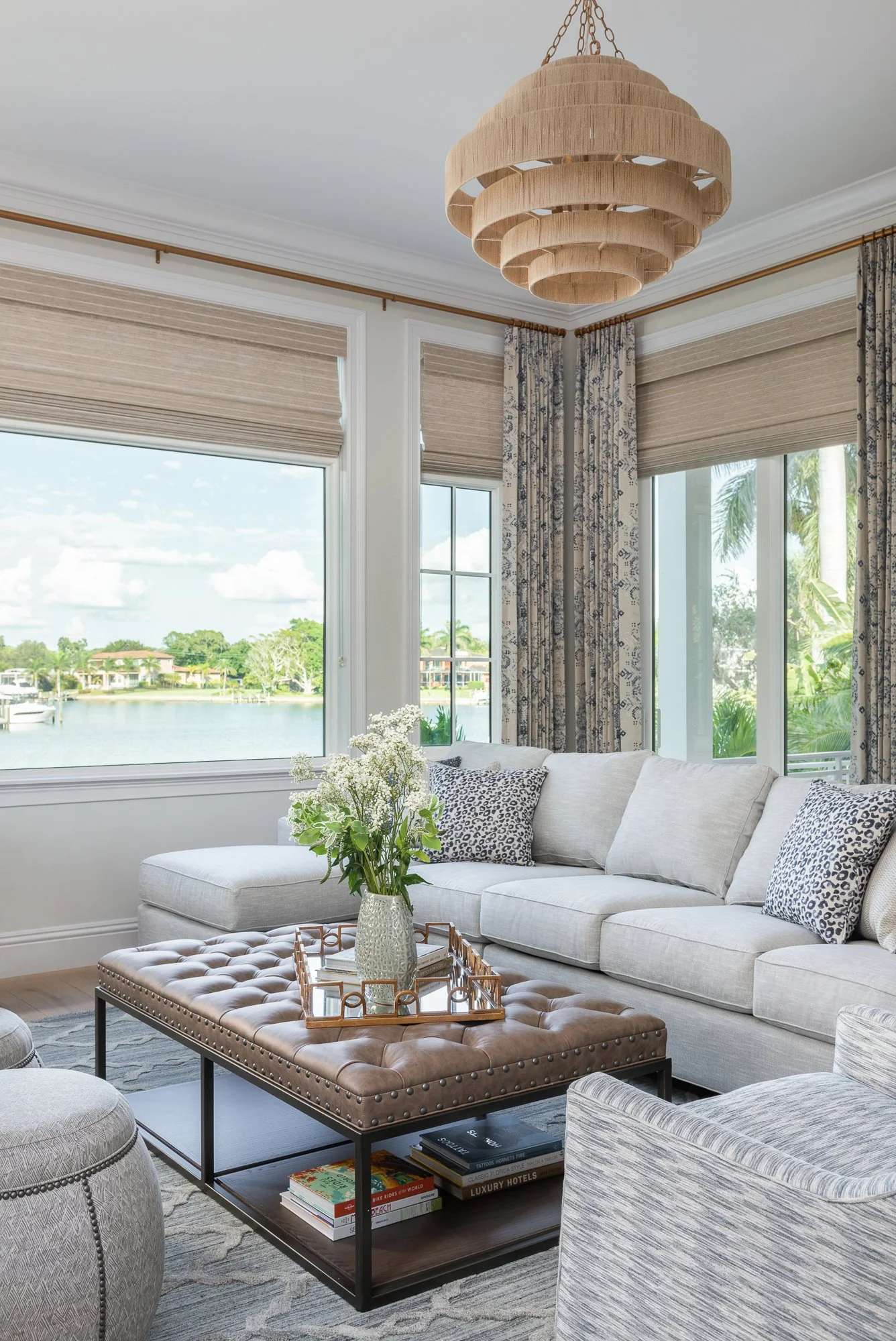 Living room with large windows overlooking a water body, decorated with patterned curtains, a white sofa with animal print pillows, a tufted leather ottoman with a vase of white flowers, and a beige modern chandelier.