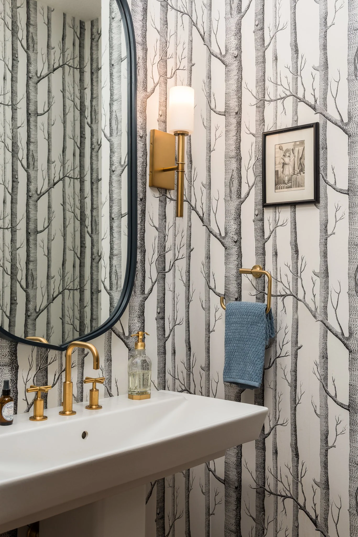 A bathroom with wallpaper featuring a black and white tree pattern, oval mirror, white sink with brass faucet, wall-mounted light fixture, framed picture, and blue towel hanging on a gold towel ring.
