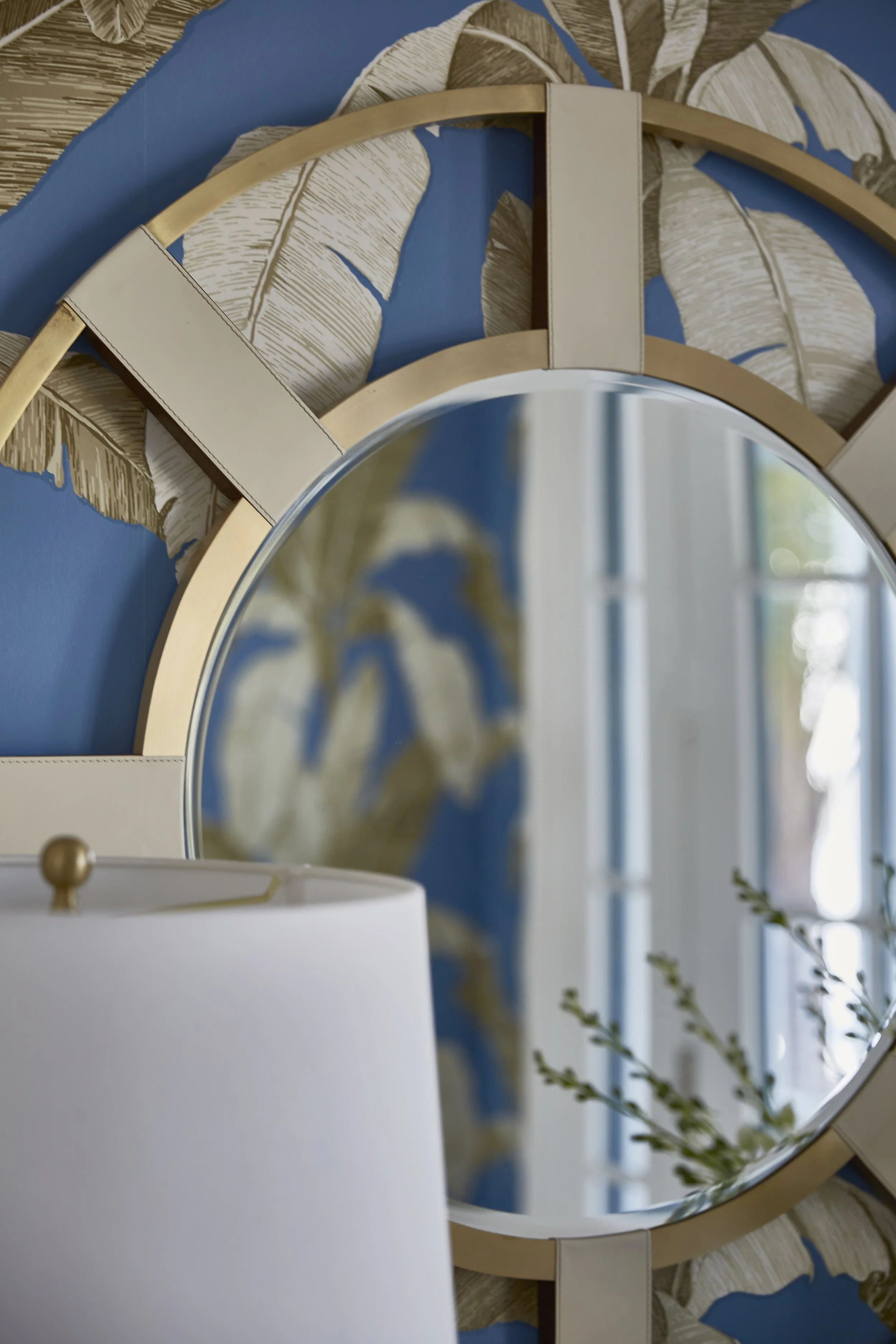 Decorative wall mirror with Ottoman-style frame, set against a blue wall with banana leaf wallpaper pattern, partial view of a white lampshade in the foreground.