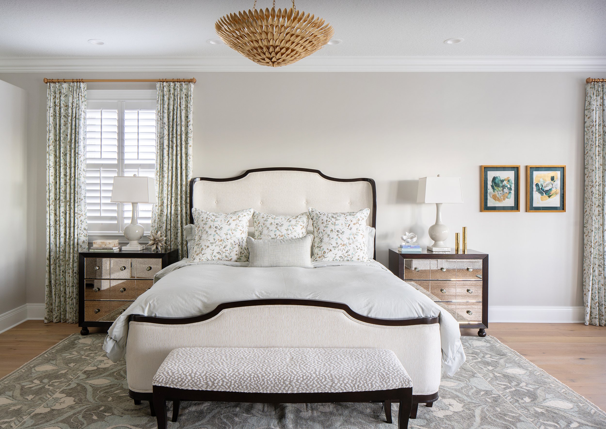 A bedroom with a white upholstered bed, bedside tables with lamps, and patterned curtains, framed abstract artwork on the wall, and a woven ceiling light fixture.