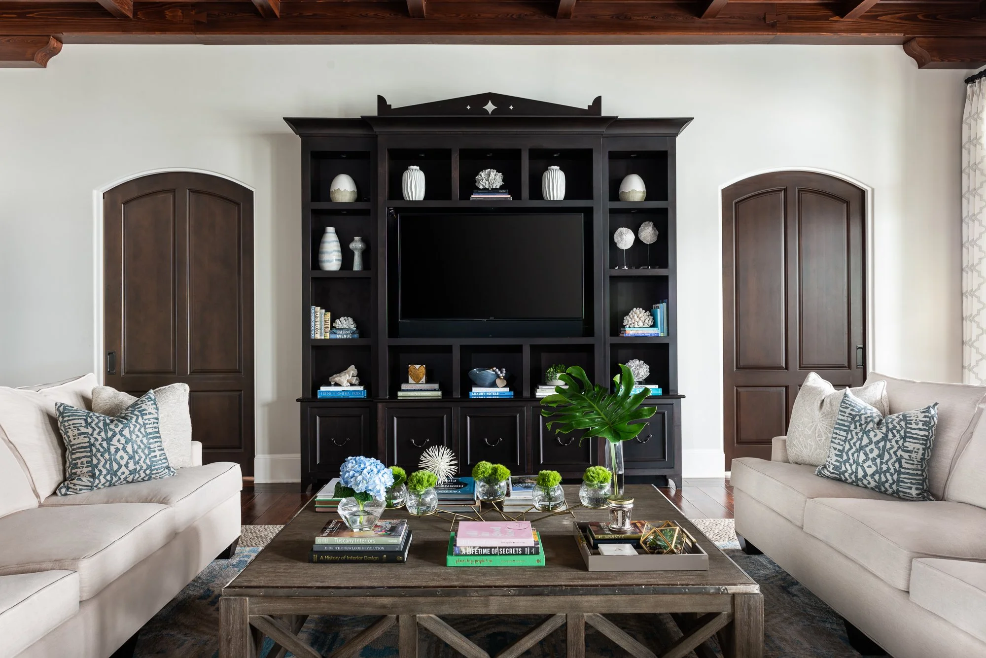 Living room with two beige sofas, a wooden coffee table with books, flowers, and decorative items, a large dark wood entertainment center with a TV, and two arched wooden doors on a white wall.
