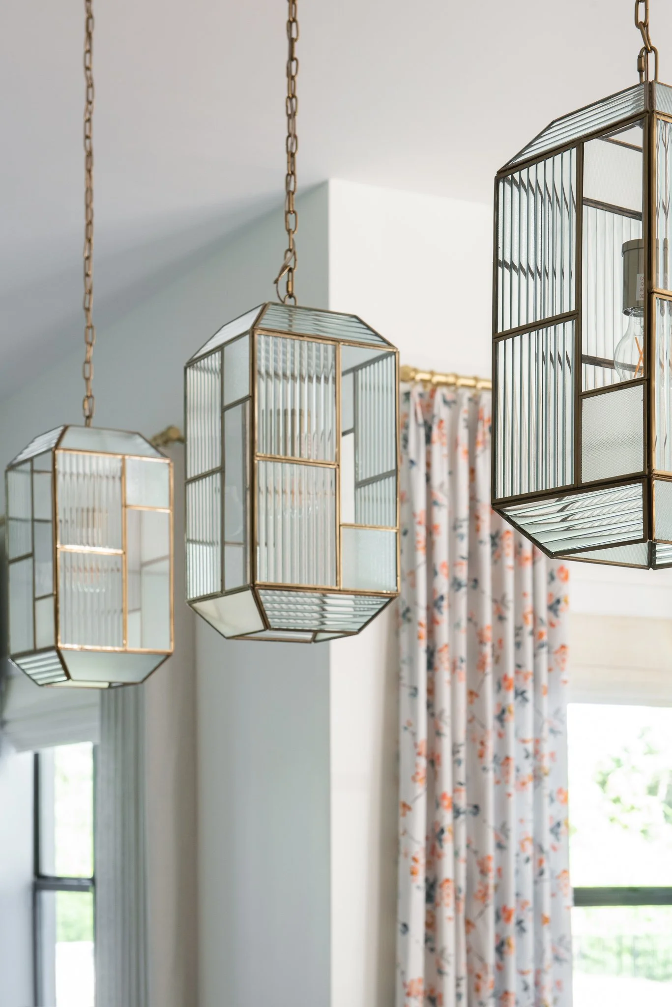 Three geometric pendant lights hanging from the ceiling in a room with floral curtains on a window.